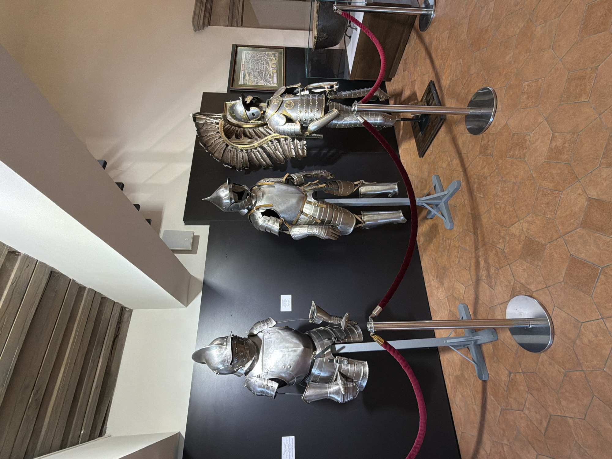 In a museum setting, three suits of ornate, polished metal armor are displayed on stands against a dark backdrop. The armor pieces appear to be full body suits, each with distinct helmet designs and detailing on the chest plates, pauldrons, and limb coverings. The leftmost suit has a more angular helmet, while the middle suit features a rounded helmet. The rightmost suit is distinguished by a large, feathered headdress attached to its helmet. Red velvet ropes on stanchions guide visitors around the exhibits. The floor is tiled with hexagonal terracotta tiles. In the background, a framed map and a large dark cauldron are visible. The ceiling features exposed wooden beams.