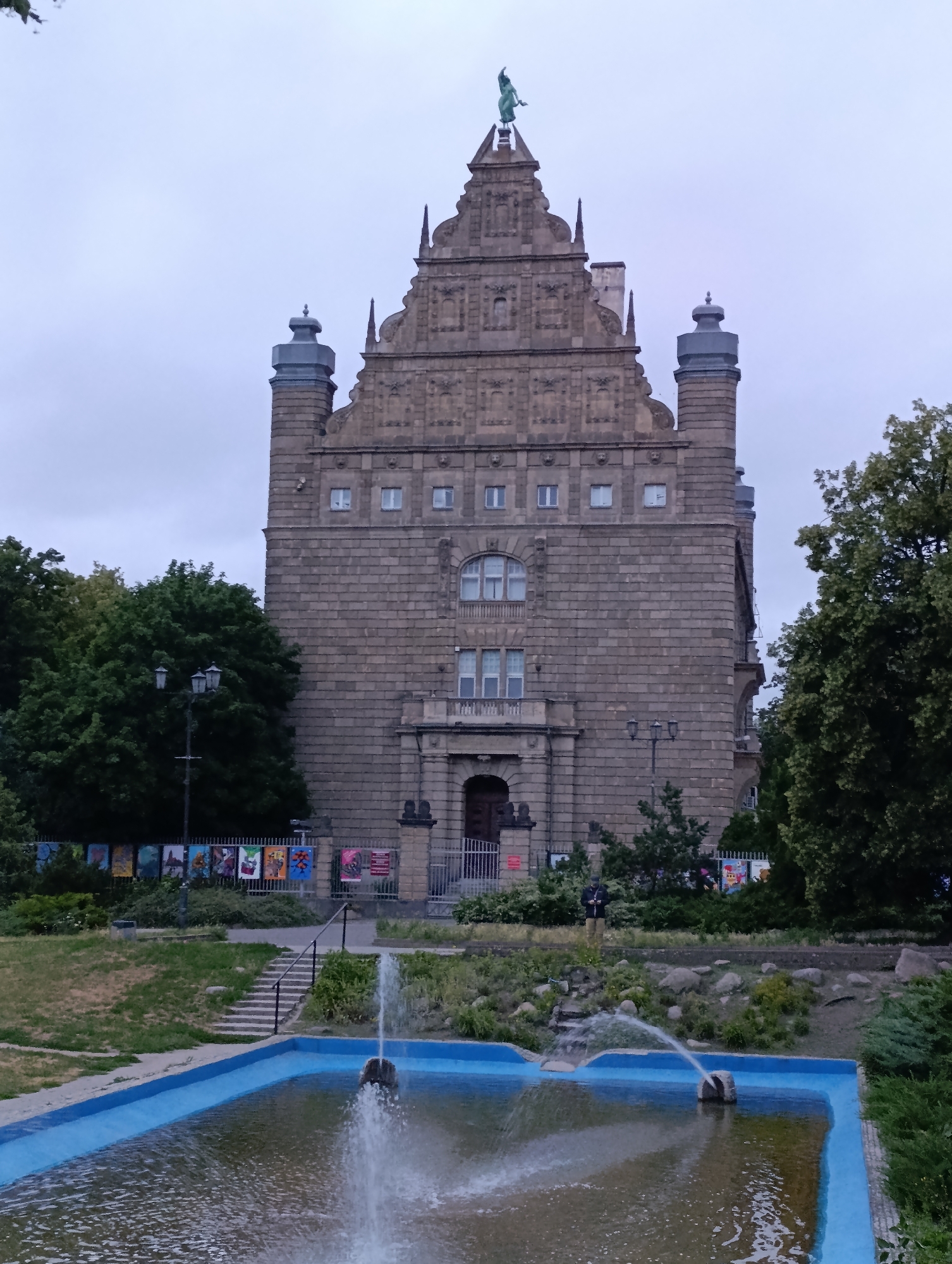 The image depicts the Artus Court in Toruń, Poland, a historic building located in the city center. The building is constructed of brick and stone, with a tall, ornate facade featuring multiple levels and a distinctive stepped gable. A statue of a figure in a green patina is visible at the very top of the building. In front of the building, there is a public square with a fountain in the foreground. The water from the fountain is spraying outwards, creating a dynamic visual effect. Surrounding the fountain are some rocks and landscaping. A set of stairs leads up from the fountain area towards the building. The area also features trees, streetlights, and a fence displaying various posters or artworks. The sky is overcast, suggesting a cloudy day. There are a few people visible in the background, one standing near the fence and another near the entrance of the building.