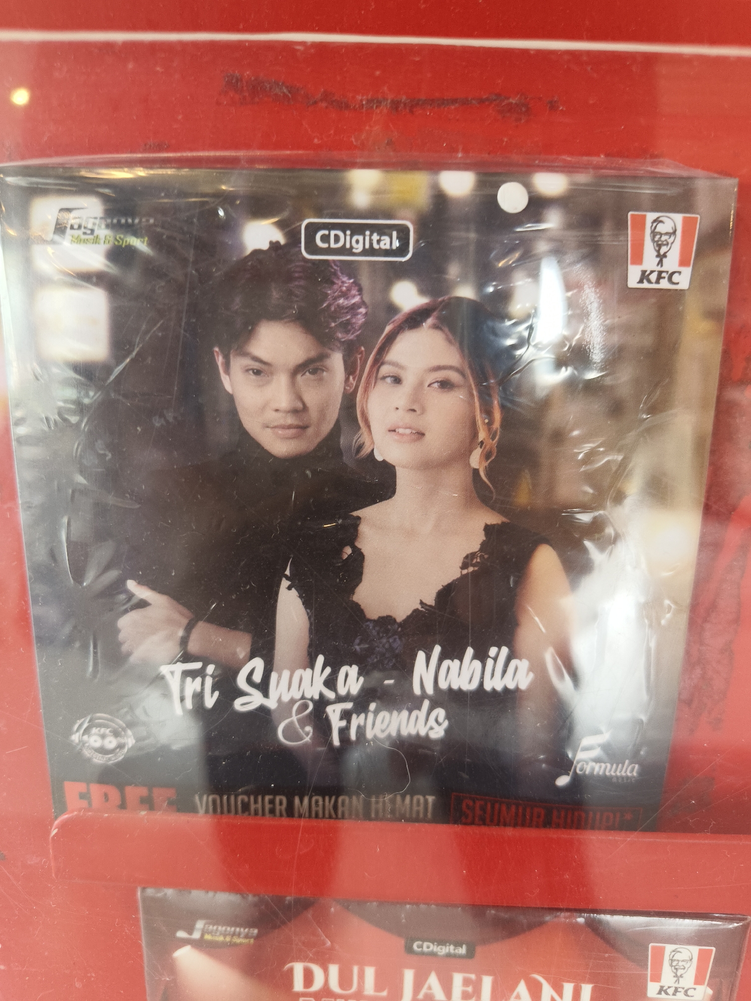 The image shows the cover of a CD featuring Indonesian artists Tri Suaka and Nabila. The CD is encased in plastic and displays the logos of "CDigital," "Formula Music," and "KFC." The text "FREE VOUCHER MAKAN HEMAT SEUMUR HIDUP" is visible at the bottom of the CD cover, indicating a promotion for a lifetime food voucher. The CD appears to be for sale or as part of a promotional package in Denpasar, Indonesia, likely within a retail environment such as a supermarket or convenience store, given the presence of other similar packaged items below. The red background suggests a display rack or shelf. The lighting is artificial, and the plastic casing of the CD creates some glare and reflections.