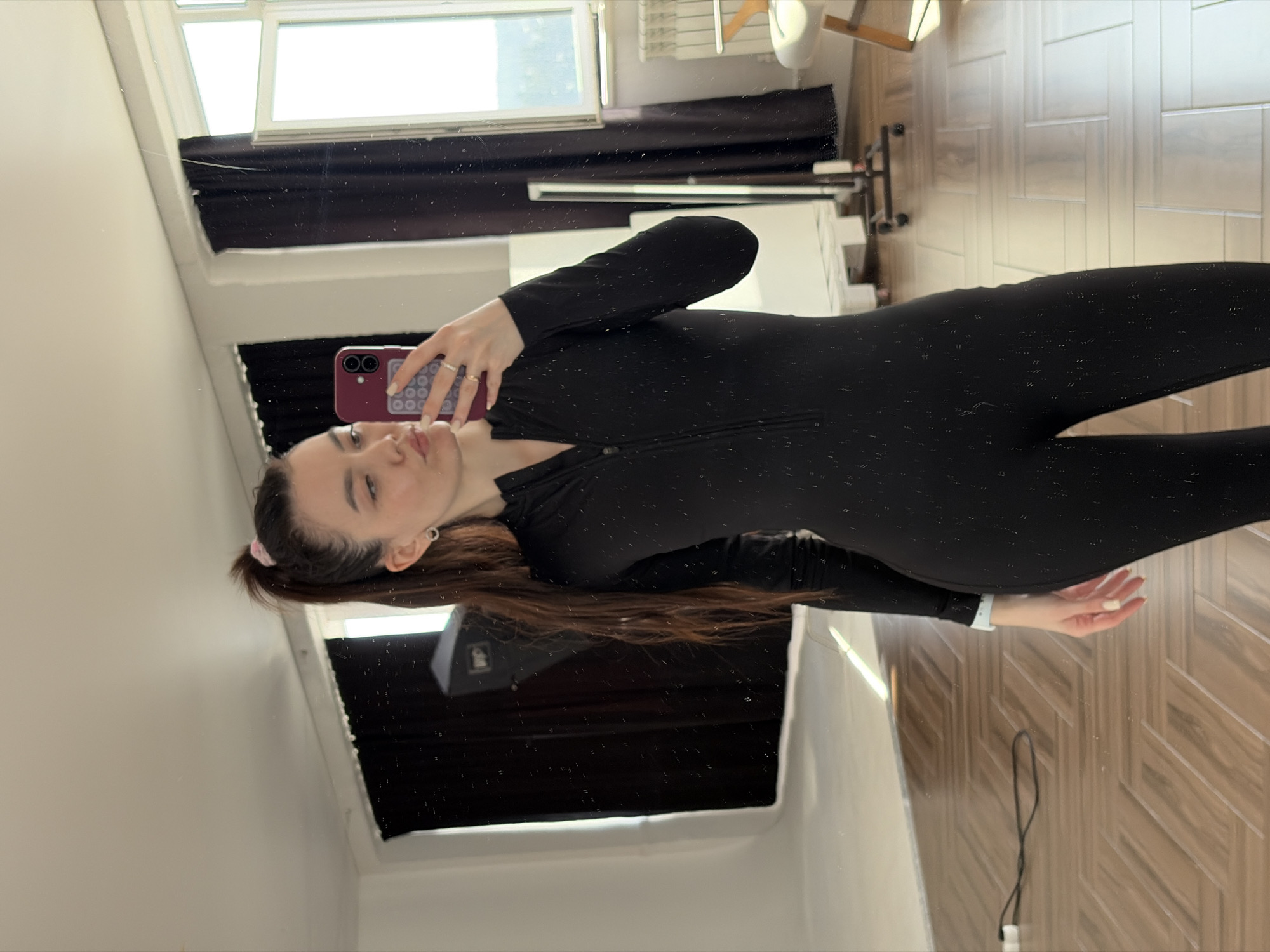 A young woman in a black jumpsuit is taking a selfie in a mirror. She is holding a purple phone with a patterned case. Her hair is brown and pulled back with a pink hair tie. The background shows a window with dark curtains, a white chair, and a parquet floor. The location appears to be inside a residence or apartment. The city of Chaikovsky, Russia, is not discernible from the image.