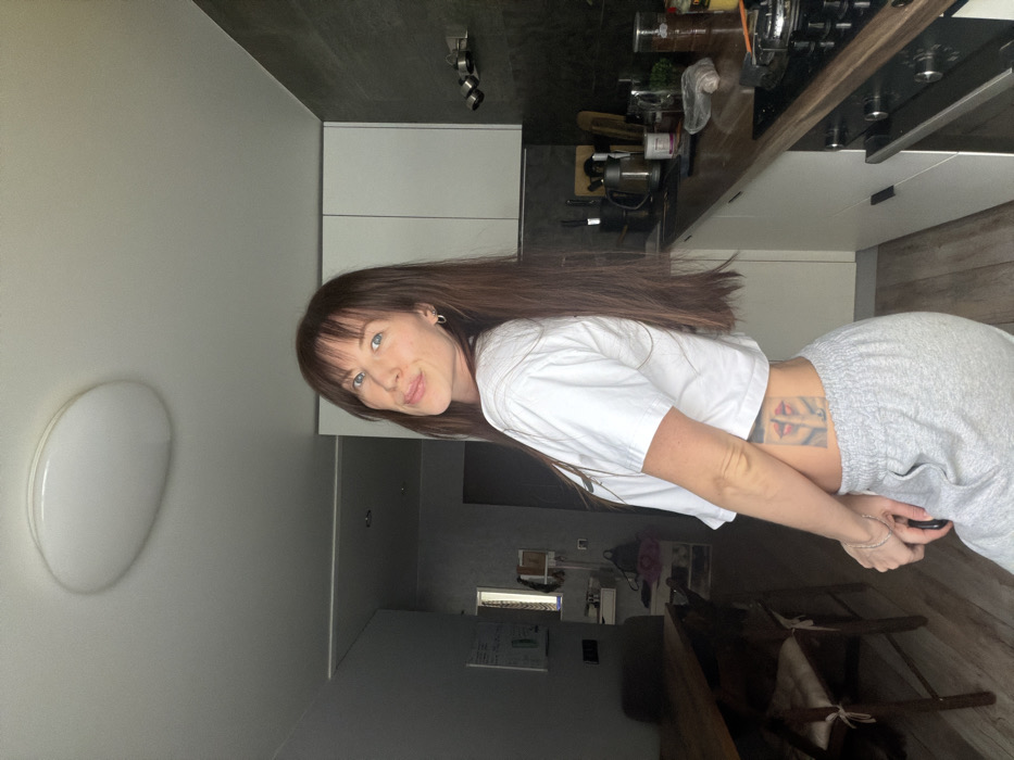 A female subject stands in an indoor residential setting in Perm, Russia, facing the camera over her left shoulder with a slight smile. She has long, dark brown hair with bangs and is wearing a white cropped t-shirt and light grey sweatpants. A tattoo depicting two sets of red lips is partially visible on her left lower torso. A thin, light-colored bracelet is present on her left wrist.
To the subject's right, a modern kitchen area is visible, featuring white upper and lower cabinets, a dark grey tiled backsplash, and a wooden-toned countertop. Objects on the countertop include a black electric kettle, a knife block, a cutting board, transparent jars containing various items (such as coffee beans and spices), a small potted plant, and a crumpled clear plastic bag. A black stovetop with two visible burners and an integrated oven are installed. A three-spotlight fixture is mounted above the countertop.
To the subject's left, an adjacent dining or living area is present, furnished with a dark wooden dining table and at least two chairs, each equipped with a light-colored cushion. The walls in this area are light grey. A white rectangular whiteboard with handwritten notes is affixed to the wall. Overhead, a white, oval-shaped flush-mount ceiling light fixture is centrally positioned.