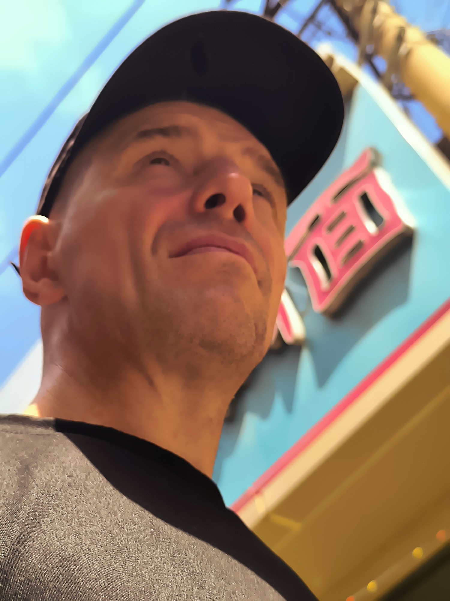 A man wearing a black baseball cap and a black t-shirt is looking up and to the right. He has short hair and is smiling. Behind him, a blue building with pink and white signage is visible. The signage appears to be in Chinese characters. The sky is bright blue.