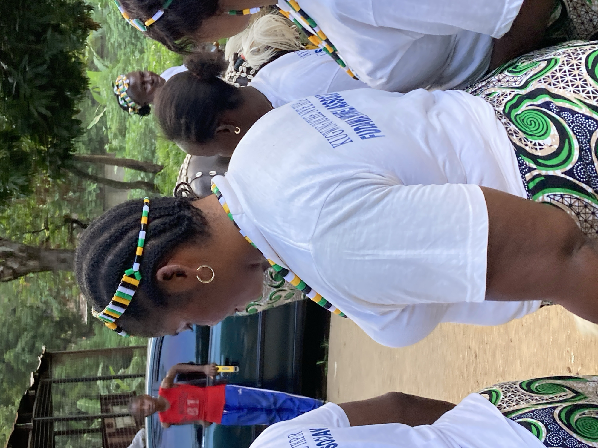 In the city of Jos, Nigeria, a group of individuals are gathered outdoors. The foreground features three women wearing white t-shirts and patterned skirts. The woman in the center, viewed from the side, has her hair braided and adorned with a colorful headband featuring green, yellow, black, and white geometric patterns. She is also wearing a gold hoop earring and a beaded necklace in similar colors. The t-shirt she is wearing has blue text on the back. To her left, another woman is partially visible, also wearing a white t-shirt and a patterned skirt. To her right, a third woman is seen from behind, wearing a white t-shirt with a colorful beaded necklace. In the mid-ground, a young boy in a red sleeveless shirt with the number "81" in white and blue pants stands near a dark-colored vehicle. Behind the people, trees and foliage create a natural backdrop, and a rustic structure with a corrugated roof is visible. The ground is a dirt surface.