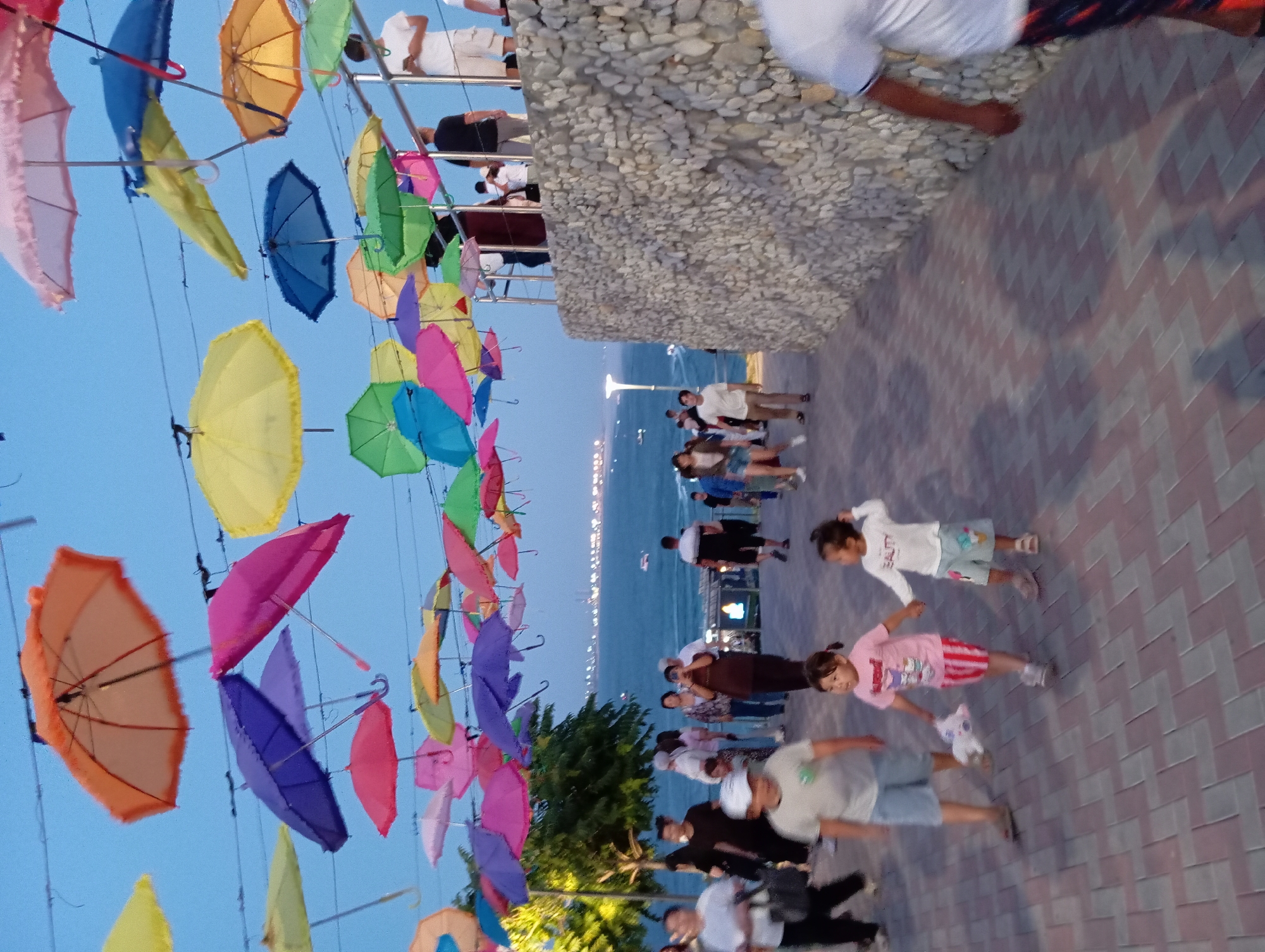 The image depicts a public walkway lined with a stone wall, adorned with numerous colorful umbrellas suspended overhead, creating a festive canopy. The walkway, paved with brick-like stones in a herringbone pattern, leads towards a body of water, likely the Caspian Sea, visible in the background. Several individuals are present, including adults and children walking along the path, some in casual attire. The scene appears to be set in the late afternoon or early evening, as indicated by the soft lighting and clear sky. This location is identified as the Aktau seaside promenade in Aktau, Kazakhstan, a popular recreational area known for its vibrant atmosphere and decorative installations.