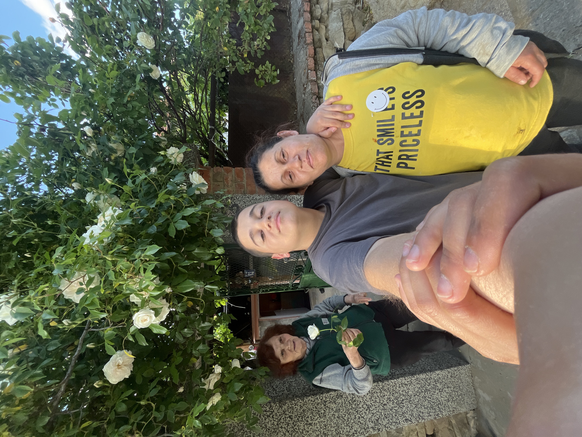 Three people are posing for a selfie in Lozarevo, Bulgaria. An older woman with short brown hair and wearing a green and gray jacket is holding a white rose. Behind her, a younger person with dark hair and a gray shirt is taking the picture. To their side, a woman with dark hair and a yellow shirt with the words "THAT SMILE IS PRICELESS" is smiling. A white rose bush is partially visible to the left. A stone wall and building are in the background. The photo appears to be taken outdoors on a sunny day.