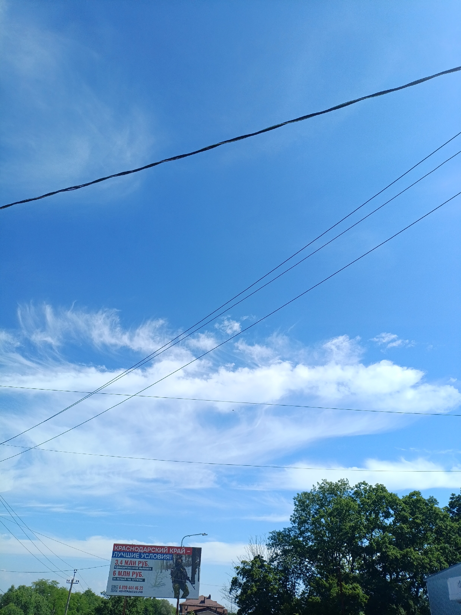 In the city of Goryachiy Klyuch, Russia, this outdoor scene captures a bright, clear blue sky with wispy cirrus clouds and scattered cumulus formations. Several power lines stretch horizontally across the frame. In the lower portion of the image, a billboard displays an advertisement for the Krasnodar Krai, featuring an image of a soldier and text in Russian indicating financial incentives for service. A tall street lamp is visible next to the billboard. Lush green trees fill the bottom right and left sides of the frame, with some buildings and other vegetation in the distance. The overall impression is a sunny day in a semi-urban or rural setting.