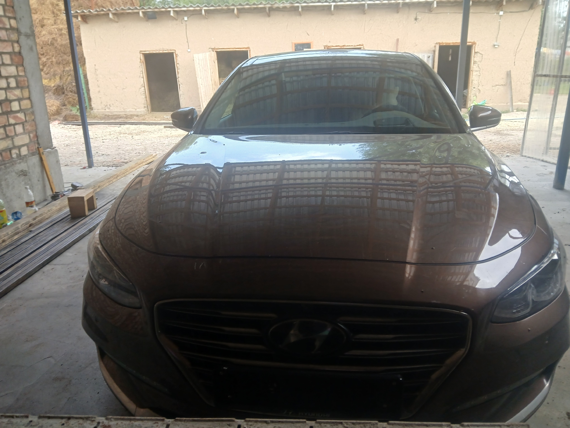 The image shows a brown Hyundai sedan parked in what appears to be a rural or semi-developed area. The car is facing the camera, and its hood and front grille are prominent. The background shows a beige building with open doorways and a roofline that suggests it might be under construction or in a state of disrepair. To the left of the car, there's a brick wall, and to the right, a clear plastic-covered structure. The ground is a mix of concrete and dirt. Several metal beams are visible stacked horizontally on the left side of the frame, and a wooden block is also present. A bottle of liquid can be seen on the left. The overall impression is an outdoor parking area, possibly a workshop or storage space, in a less urbanized setting. The location is identified as Mihaylovka, Kyrgyzstan.