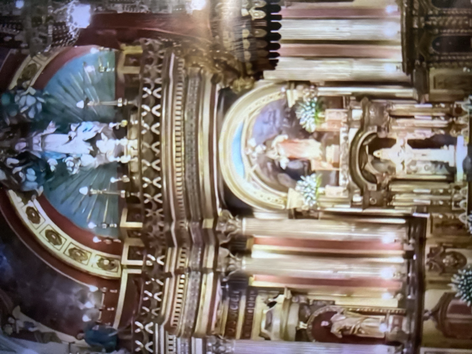 The image shows a baroque style altar piece, likely within a church or cathedral. The location is suggested to be Tinaquillo, Venezuela. The altar piece is richly decorated with gold and ornate carvings. There are figures of saints or religious figures displayed prominently. There are multiple levels to the structure, and columns and architectural details characteristic of the baroque style are evident. The lighting appears dim and perhaps natural light illuminates it.