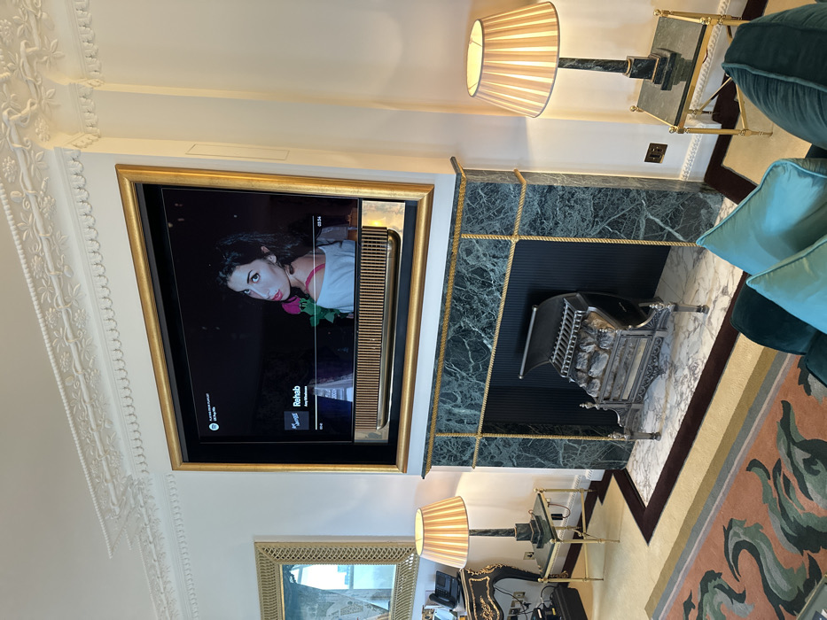The image depicts an opulent interior, likely a luxury hotel suite, located in Central London, United Kingdom. A large television screen, framed in gold, is centrally mounted above a dark green marble fireplace. The screen displays the Spotify interface, playing the song "Rehab" by Amy Winehouse, with her image featured prominently.
The fireplace features a green marble mantelpiece with a gold rope-textured trim, a black fluted firebox interior, and a cast-iron grate containing artificial logs, resting on a white marble hearth.
Flanking the fireplace are two identical side tables with gold-toned legs and green marble tops. Each table holds a striped table lamp with a cream-colored pleated shade and a dark marble base.
The walls are white, adorned with elaborate white plaster crown molding featuring intricate floral and classical motifs. A large gold-framed mirror is visible on the wall to the left, reflecting an outdoor scene. Below the mirror, a dark, rococo-style table holds a black office phone and other electronic devices.
In the foreground, a teal velvet sofa with light blue cushions is partially visible. The floor features a light-colored carpet with a dark border, partially covered by a large rug with an abstract pattern in shades of teal, green, and terracotta. The overall ambiance is one of classic luxury and grandeur.