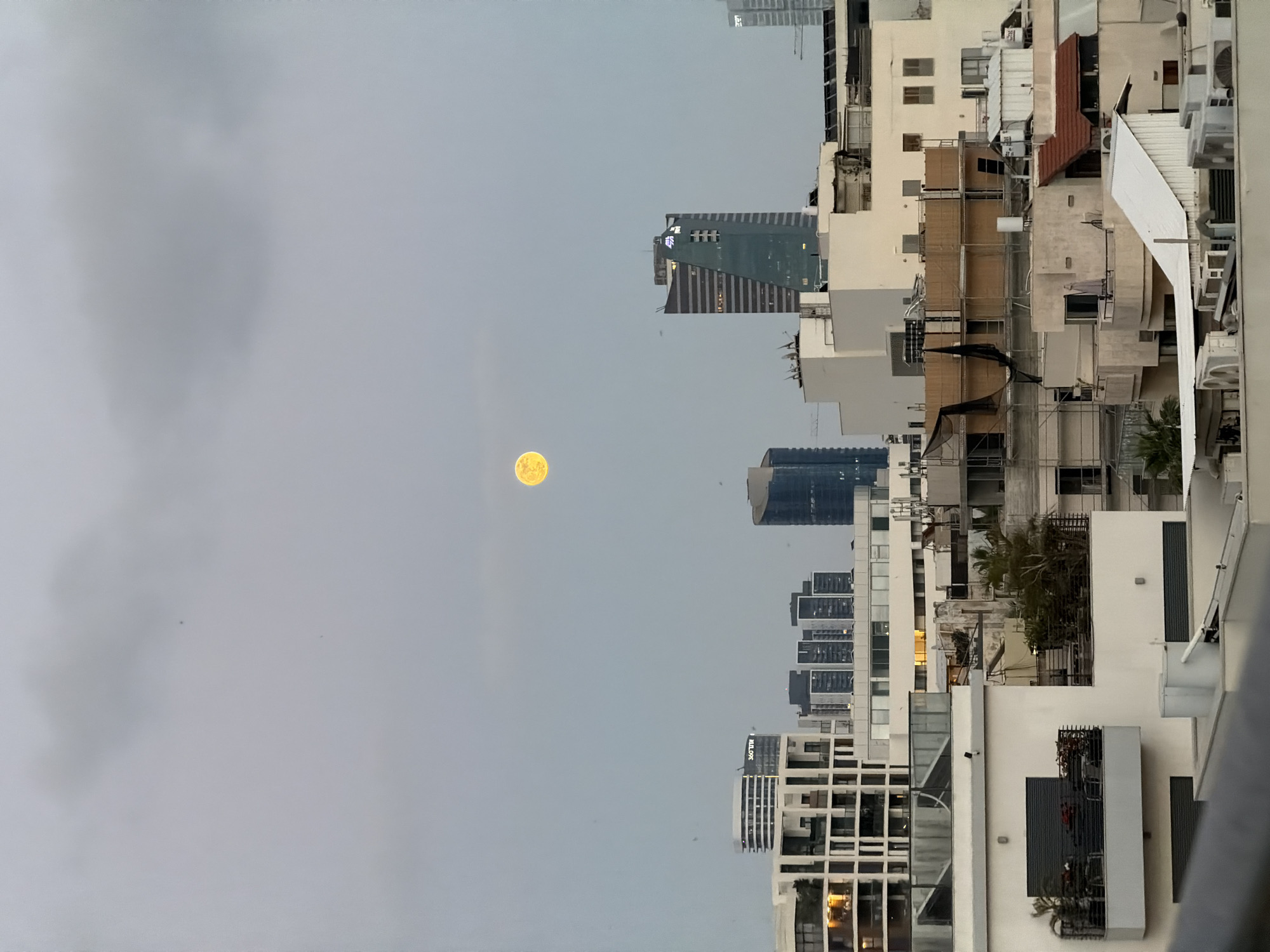 The image shows a nighttime view of the city of Tel Aviv, Israel. The sky is overcast, with a full moon visible through the clouds. Several high-rise buildings are visible, as well as older residential buildings. The buildings are a mix of modern and older architecture. The photograph appears to have been taken from a high vantage point, such as a balcony or rooftop. The name "360TLB" is visible on a high-rise building.