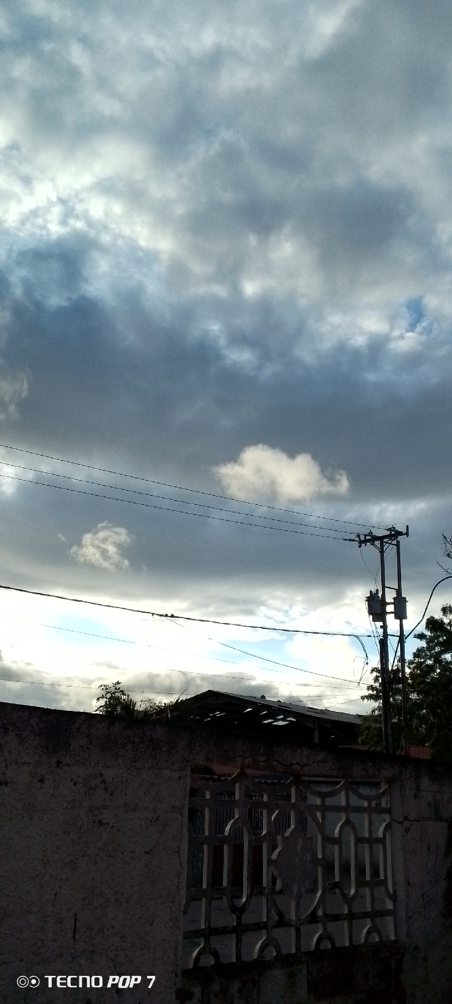 The image shows an overcast sky over a residential area in Altagracia, Venezuela. Heavy, dark clouds dominate the sky, with a few patches of lighter clouds. Power lines stretch across the middle of the frame, connected to a utility pole with a transformer. Below, a dilapidated building is partially visible behind a weathered concrete wall with an ornate metal grate. Some vegetation, likely trees or shrubs, appears behind the building. The lower left corner of the image displays the "TECNO POP 7" watermark.