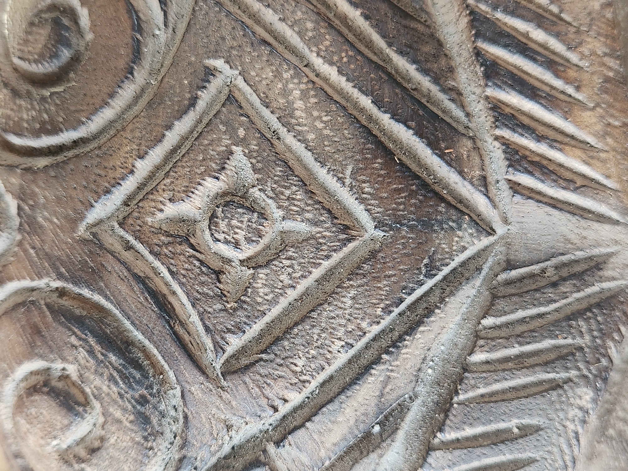 This image is a close-up, overhead shot of a carved wooden surface. The carving features a central diamond shape with an inner four-petal flower design. Surrounding the diamond are carved lines that radiate outwards and curved, swirling patterns. The wood appears aged and weathered, with visible grain and some dust or debris within the carved crevices. There is no indication of the specific location within the city of Maitland, United States of America, or any people, actions, or interactions. The image focuses solely on the texture and detail of the carving.