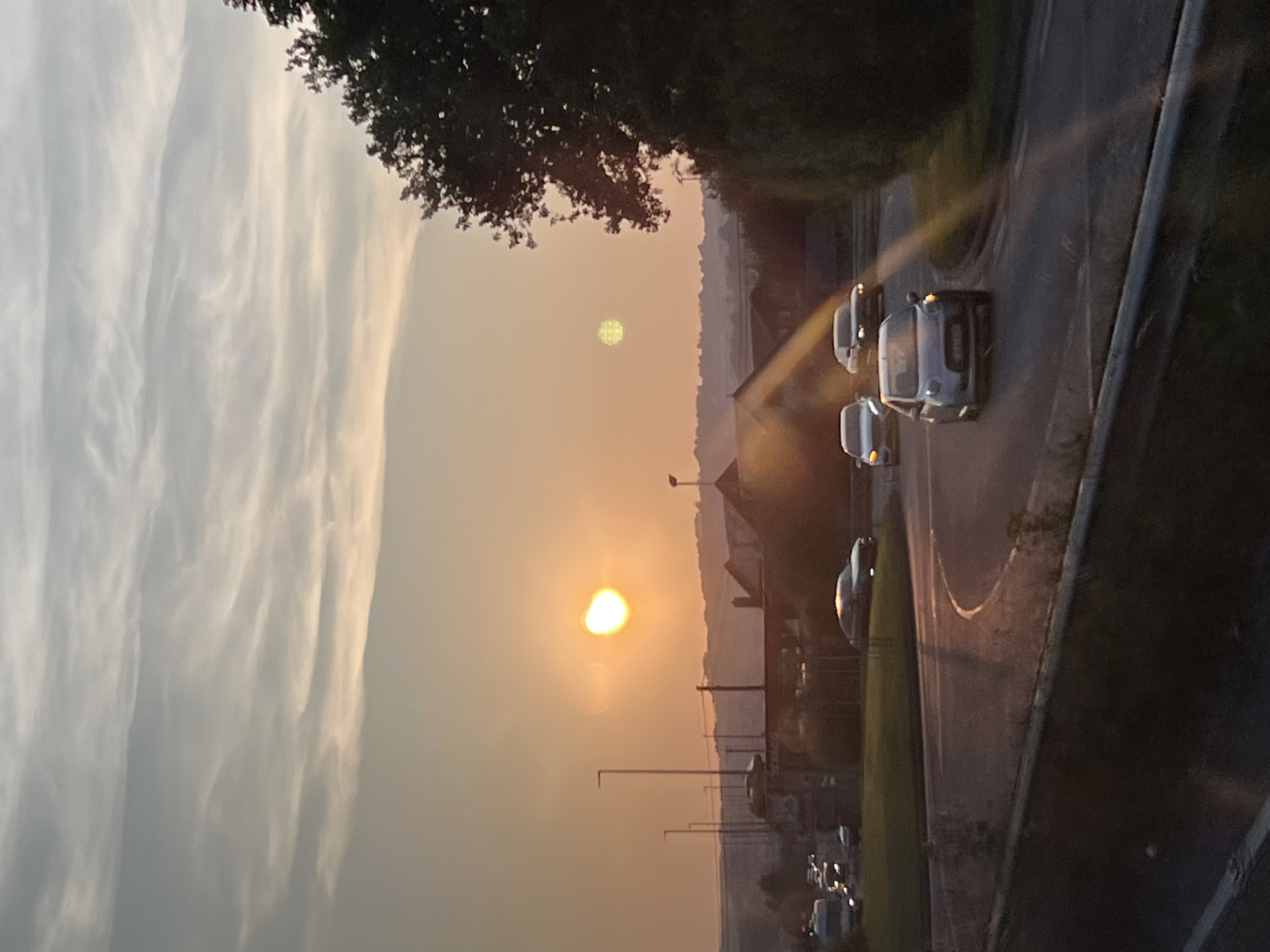 The image depicts a sunset over a road in Letterkenny, Ireland. A bright orange sun is visible in the sky, casting a warm glow on the scene. Several cars are driving on a curving road, with streetlights and buildings visible in the background. A large tree on the right side of the image frames the sunset. The sky is filled with wispy clouds, and the overall atmosphere is peaceful and serene.