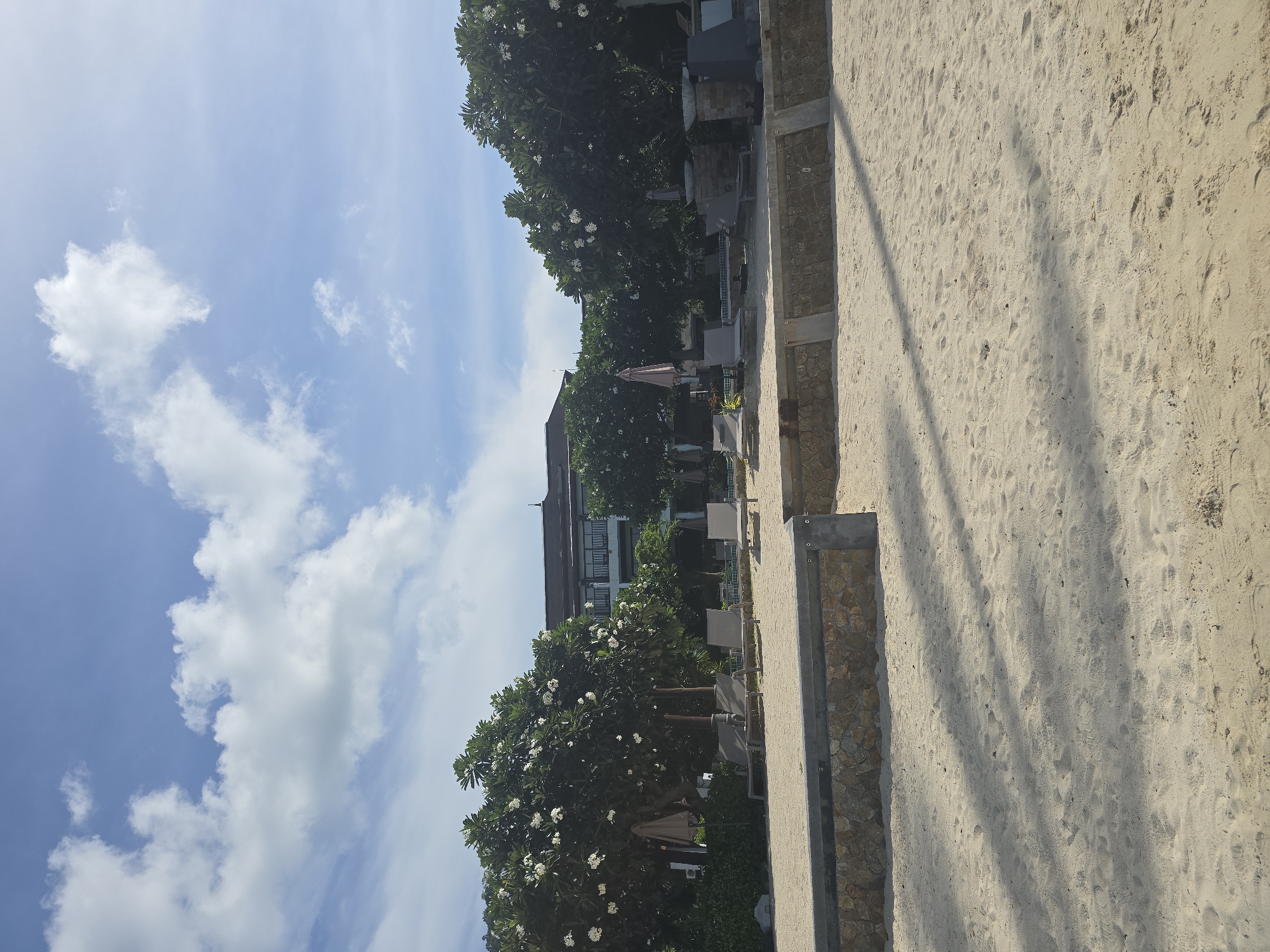 This is an outdoor, eye-level shot taken on a sandy beach in Baan Chaweng Noi, Thailand. The scene depicts a resort area with a building in the background, flanked by lush green trees with white flowers. In the foreground, there are several lounge chairs on the sand, suggesting a relaxing atmosphere. The sky above is a clear blue with scattered white clouds. The sand is lightly textured with shadows cast from unseen objects.