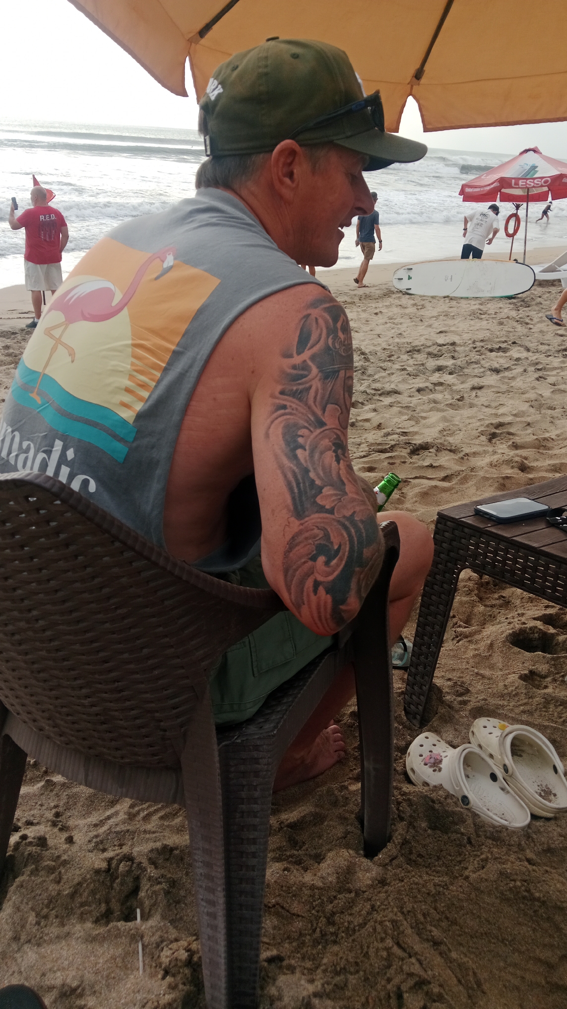 A man wearing a grey tank top with a flamingo design and green shorts sits in a brown plastic chair on a sandy beach. He has a tattoo on his right arm. Beside him, on a small wooden table, is a green bottle and a black smartphone. Two white clogs are visible in the sand near the table. In the background, people are walking along the shore, and the ocean waves are visible. An orange umbrella shades the area where the man is sitting. This scene is located on Kuta Beach in Kuta, Indonesia.