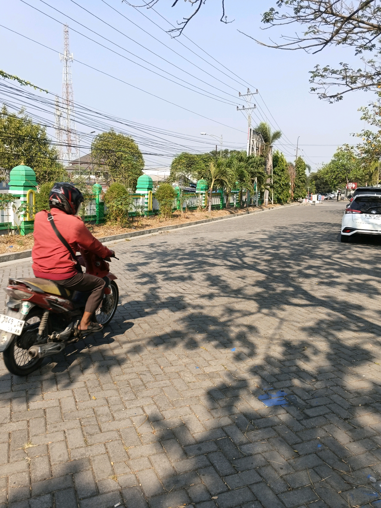 A person wearing a red jacket and a helmet is riding a red motorcycle on a paved street. The street is lined with green fences and trees. In the background, there is a tall telecommunications tower and a clear blue sky. The scene appears to be in an urban or suburban environment, likely in Semarang, Indonesia, given the style of the motorcycle's license plate and the surrounding architecture. The sun is shining, casting shadows of trees onto the street. A white car is parked on the right side of the street.