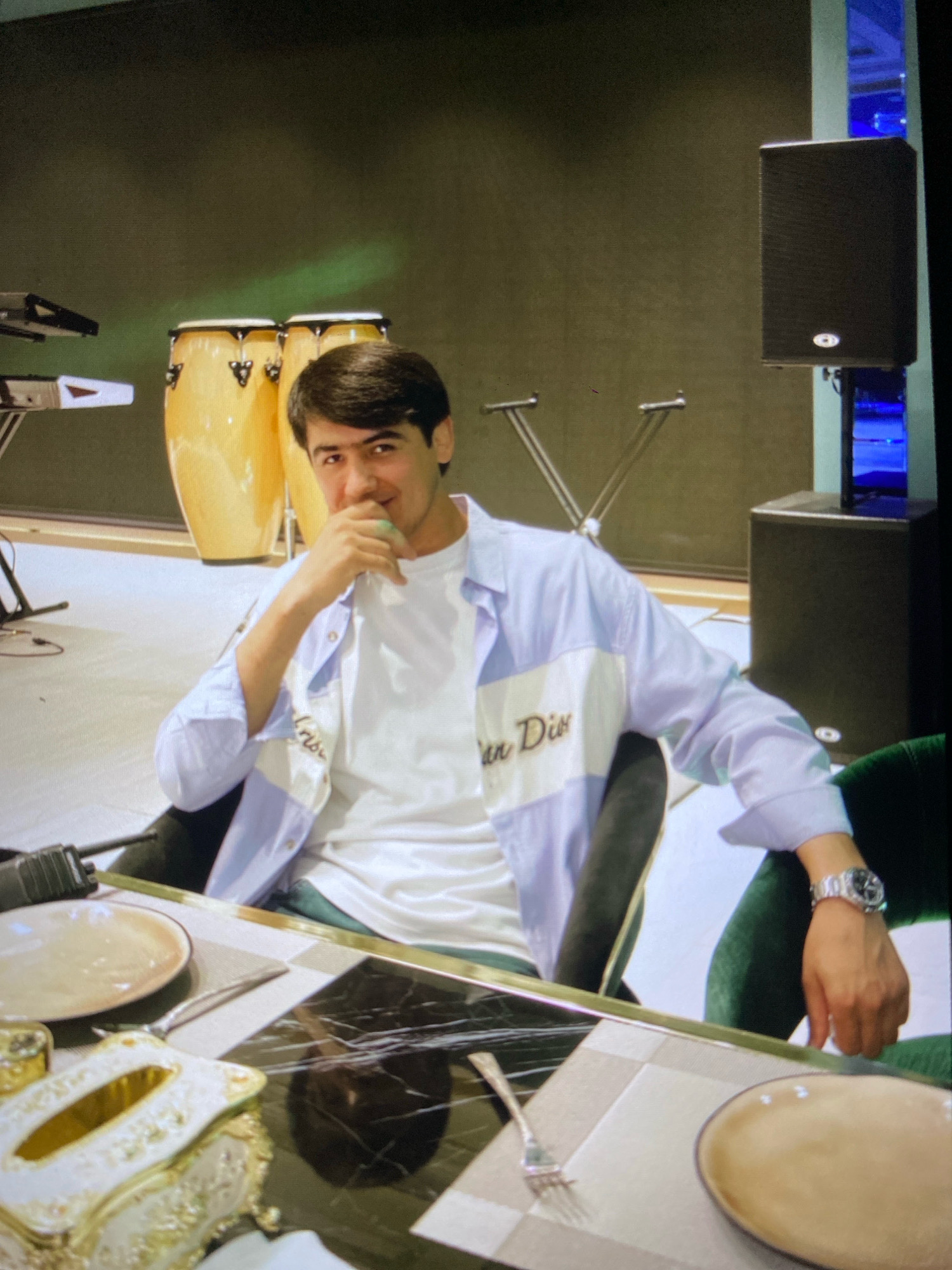 A young man with dark hair and a light complexion is seated at a table, looking towards the camera. He is wearing a white t-shirt and an open light blue collared shirt. His right hand is raised to his chin, and he is smiling. The table is set with plates and cutlery. Behind him, there are two large yellow conga drums and part of a keyboard on the left. On the right, a large black speaker is mounted on a stand. The background is a dark, textured wall. The lighting appears to be artificial and somewhat dim. The scene is indoors, in what could be a studio or a venue with musical equipment. There is no discernible indication of the location being in the city of Ramenskoye, Russia.