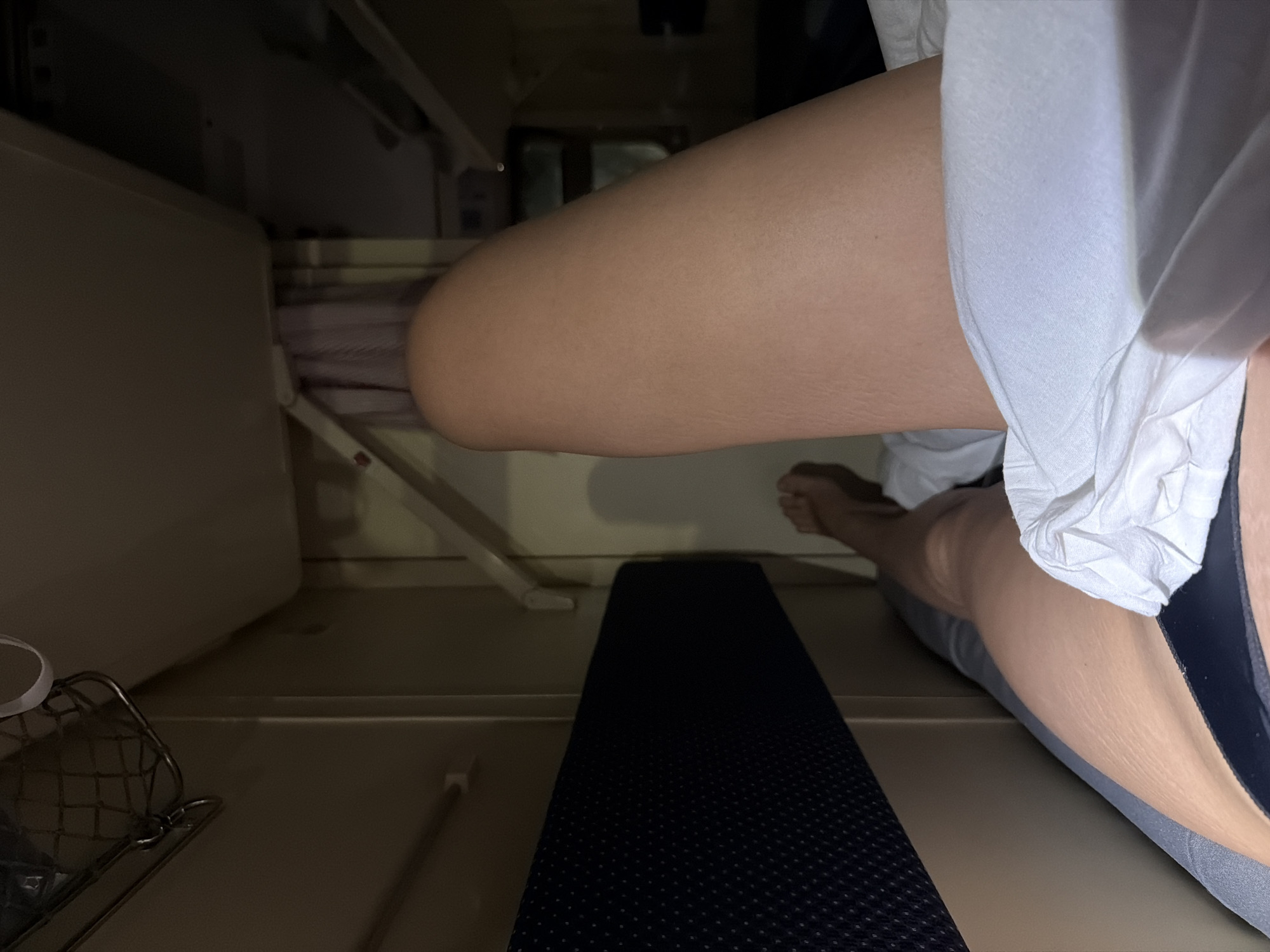 A low-angle, close-up shot shows a person sitting within a confined space. A fair-skinned thigh and leg, partially covered by white fabric (possibly a shirt or underwear), are visible. The person's foot is visible. A dark, rectangular object, possibly a mat or cushion, lies on the surface the person is sitting on. Parts of metal rack or shelving can be seen. The environment is dimly lit. The image does not contain enough information to establish the location as Октябрьский, Russia.