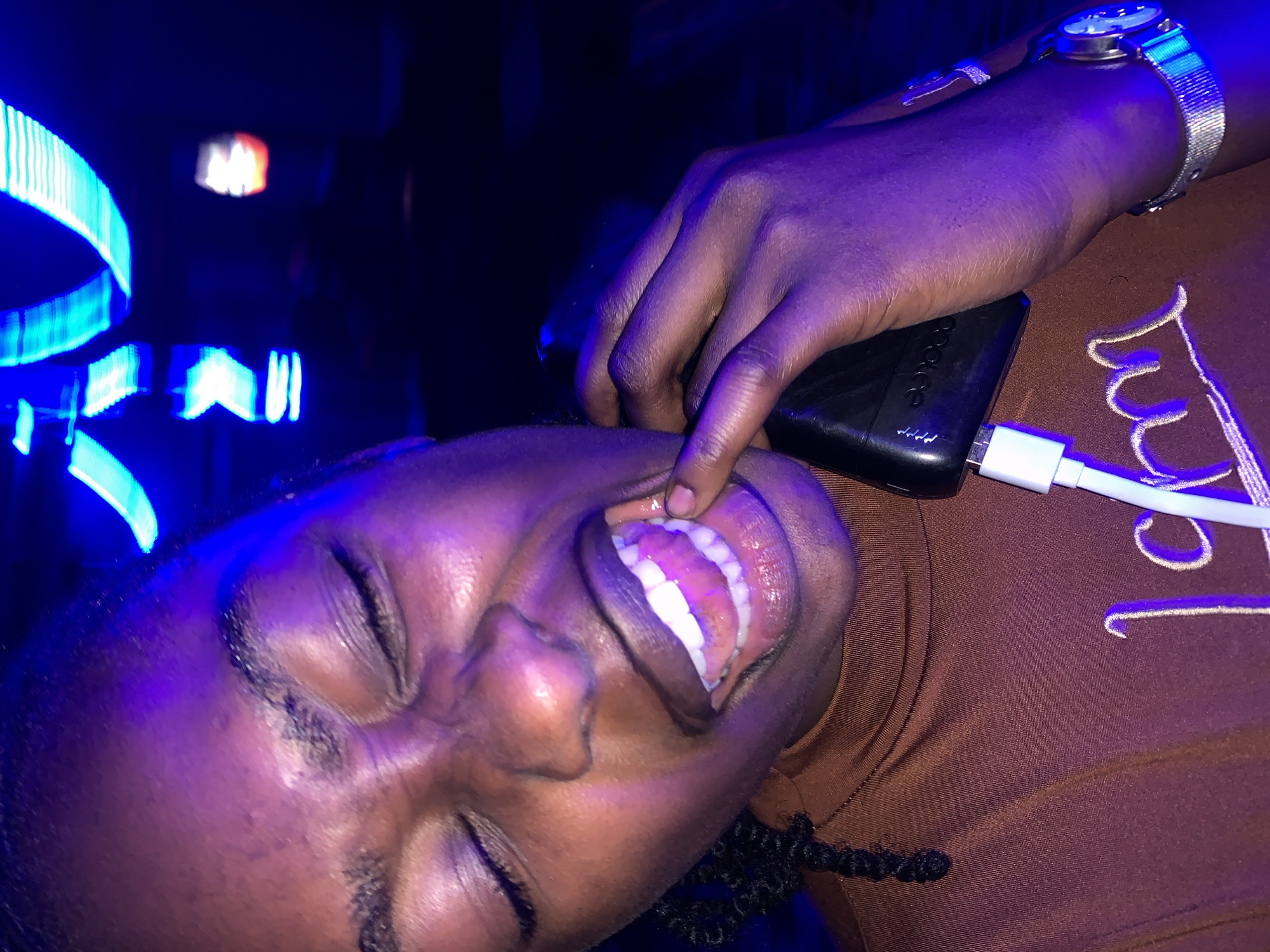 The image depicts an individual in the city of New Stadium, Nigeria, captured under prominent blue and purple ambient lighting. The person, appearing to be an adult with dark skin, has their eyes closed and their face is contorted in an expression of intense emotion. The mouth is open wide, with a finger from the left hand pulling down the lower lip to fully expose the lower gum line and teeth. The individual is wearing a brown, high-necked garment featuring partially visible white embroidered text that reads "I am." A black smartphone is held in the left hand, into which a white charging cable is connected. A silver-colored wristwatch with a mesh band is worn on the left wrist. The background is dark and out of focus, displaying blurred blue linear light streaks and an indistinct red-orange light source, consistent with an indoor, low-light entertainment environment.