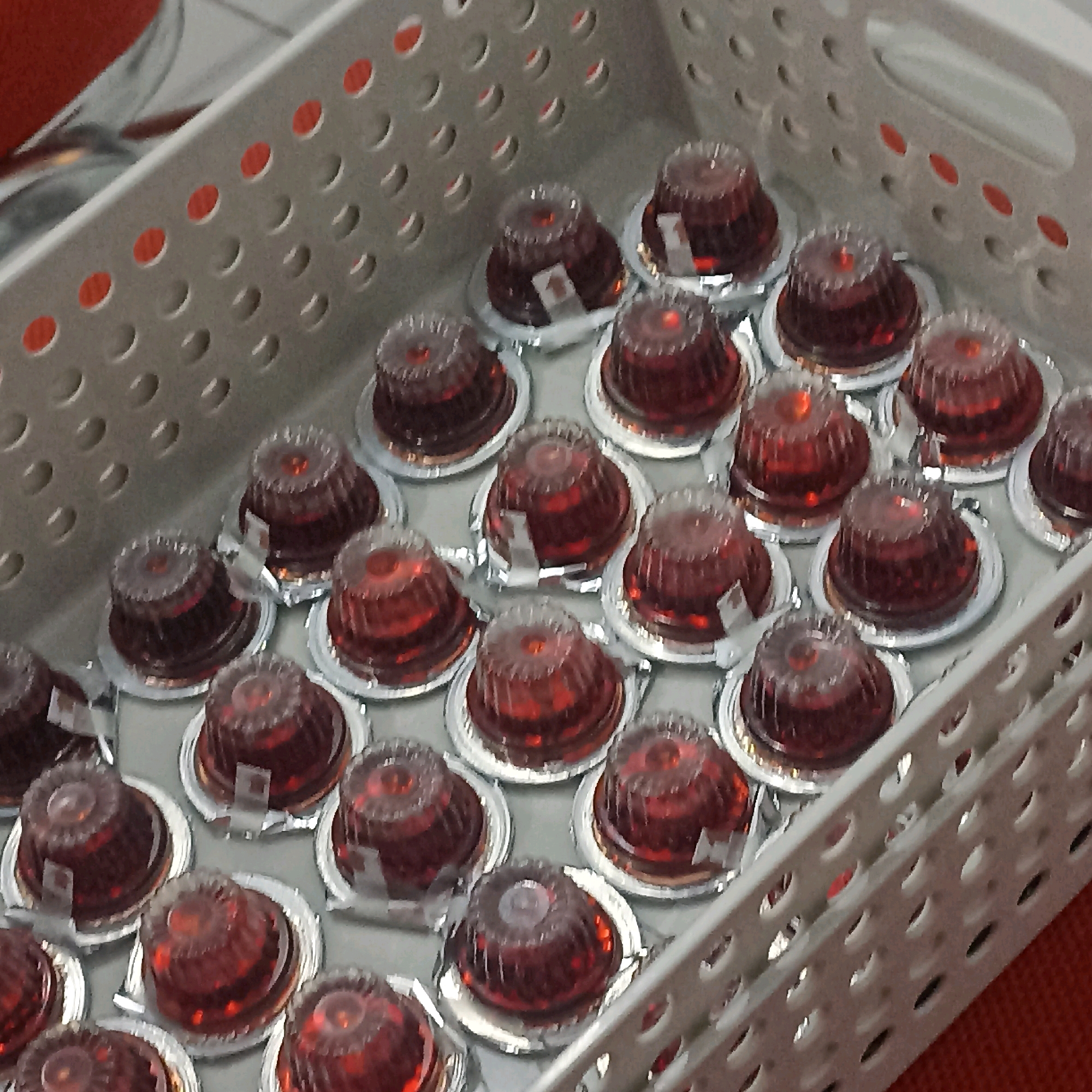 The image displays a close-up, overhead view of a white, perforated plastic basket filled with numerous small, individually-portioned red jellies. Each jelly is contained within a clear plastic cup, sealed with a silver foil lid, and sits on a silver base. The jellies are arranged in rows and columns within the basket, with the basket itself positioned on a textured orange surface. The basket has a handle visible on the right side. The lighting is somewhat dim, casting subtle shadows. There are no people visible in the image. There is no information available to determine the location in Magelang, Indonesia, or any specific context for this scene.