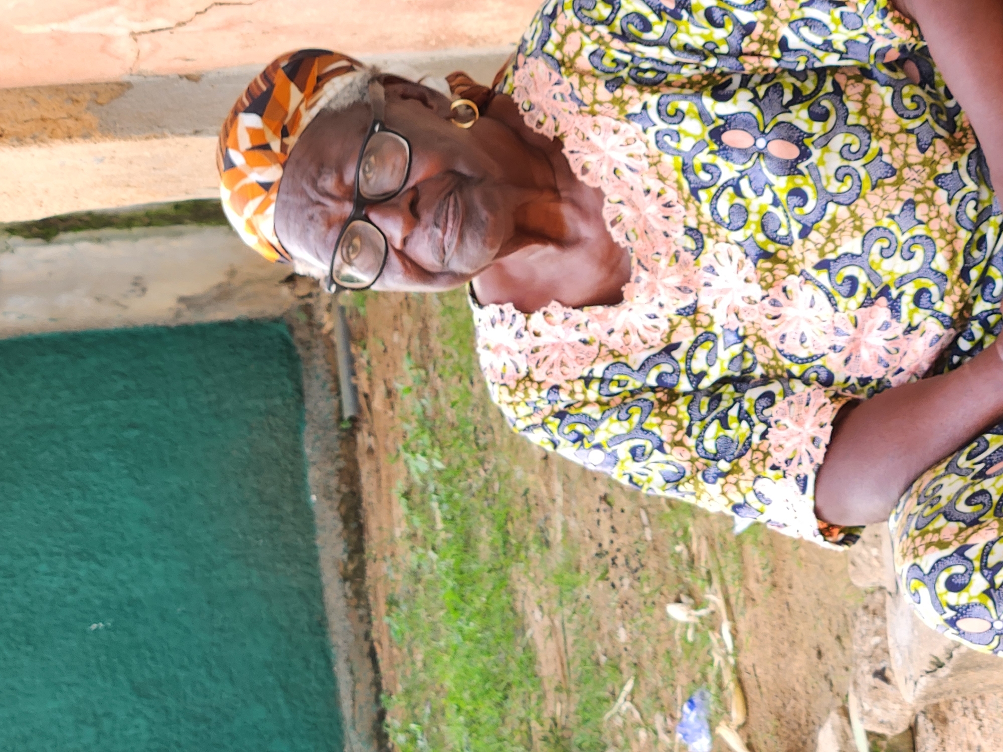 A medium shot shows an elderly Black woman sitting outdoors in Sabon Tasha, Nigeria. The woman is looking to her right, with her head slightly tilted down. She is wearing glasses, a headscarf with an orange, white, and black geometric pattern, and a colorful traditional African print dress with a pink lace trim around the neckline. A gold hoop earring is visible in her right ear. She is seated on a rough, grey stone. The background consists of a textured green wall on the left, a cracked beige wall on the right, and dry, patchy grass and dirt in the middle ground. A plastic bag is visible on the ground to the left of the woman.