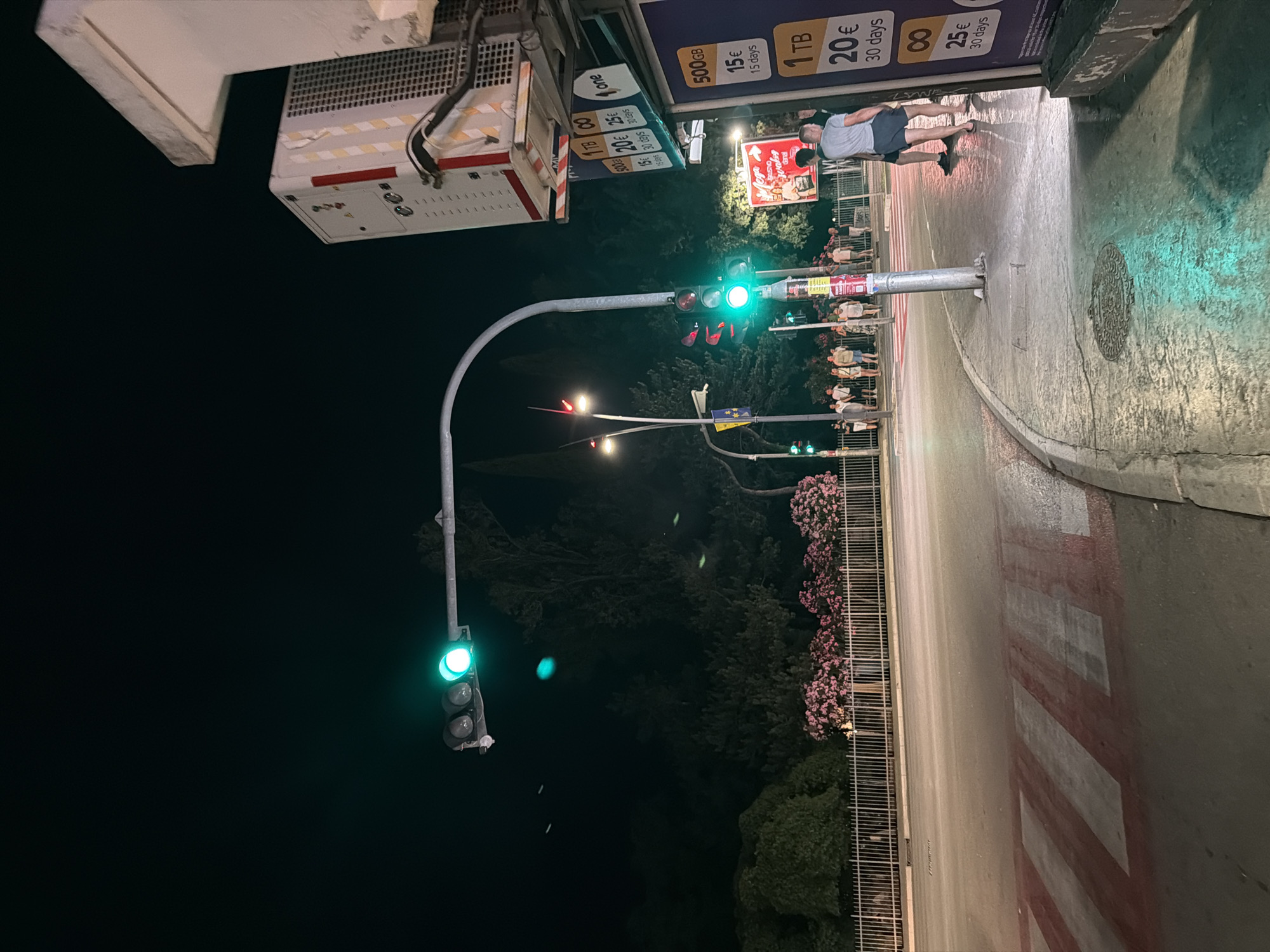 The image depicts a nighttime street scene at a pedestrian crossing in Budva, Montenegro. A traffic light is illuminated green, signaling that vehicles may proceed. Several people are gathered near the sidewalk, appearing to wait or observe. Advertisements for data plans are visible on a kiosk, offering various storage sizes and prices. The street and sidewalk are illuminated by streetlights and the glow of the traffic signal. Trees and flowering bushes line the background, suggesting a park or green space adjacent to the road. A metal fence separates the sidewalk from the green area. The overall atmosphere is one of urban nighttime activity.