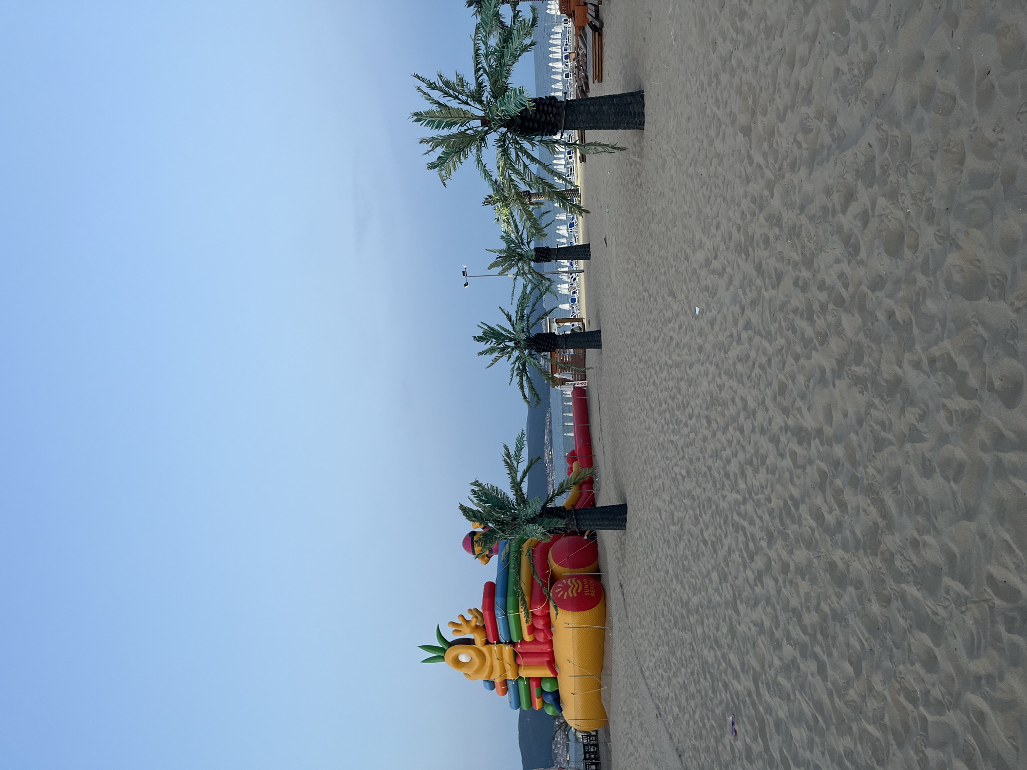 This is a daytime, horizontal, eye-level, full shot of an inflatable playground on a sandy beach in Nesebar, Bulgaria. The inflatable structure is brightly colored, with a yellow base, red and blue inflatable elements, and a pineapple-shaped character on top. A palm tree stands in front of the inflatable structure, obscuring part of it. In the background, several other palm trees line the beach, leading to a row of white umbrellas and lounge chairs. The ocean is visible in the distance, with a hazy shoreline and buildings. The sky is a pale blue, with some clouds. The sand is light brown and appears smooth, with some footprints visible. There are no people in the frame. The scene is peaceful and appears to be set up for recreational use.