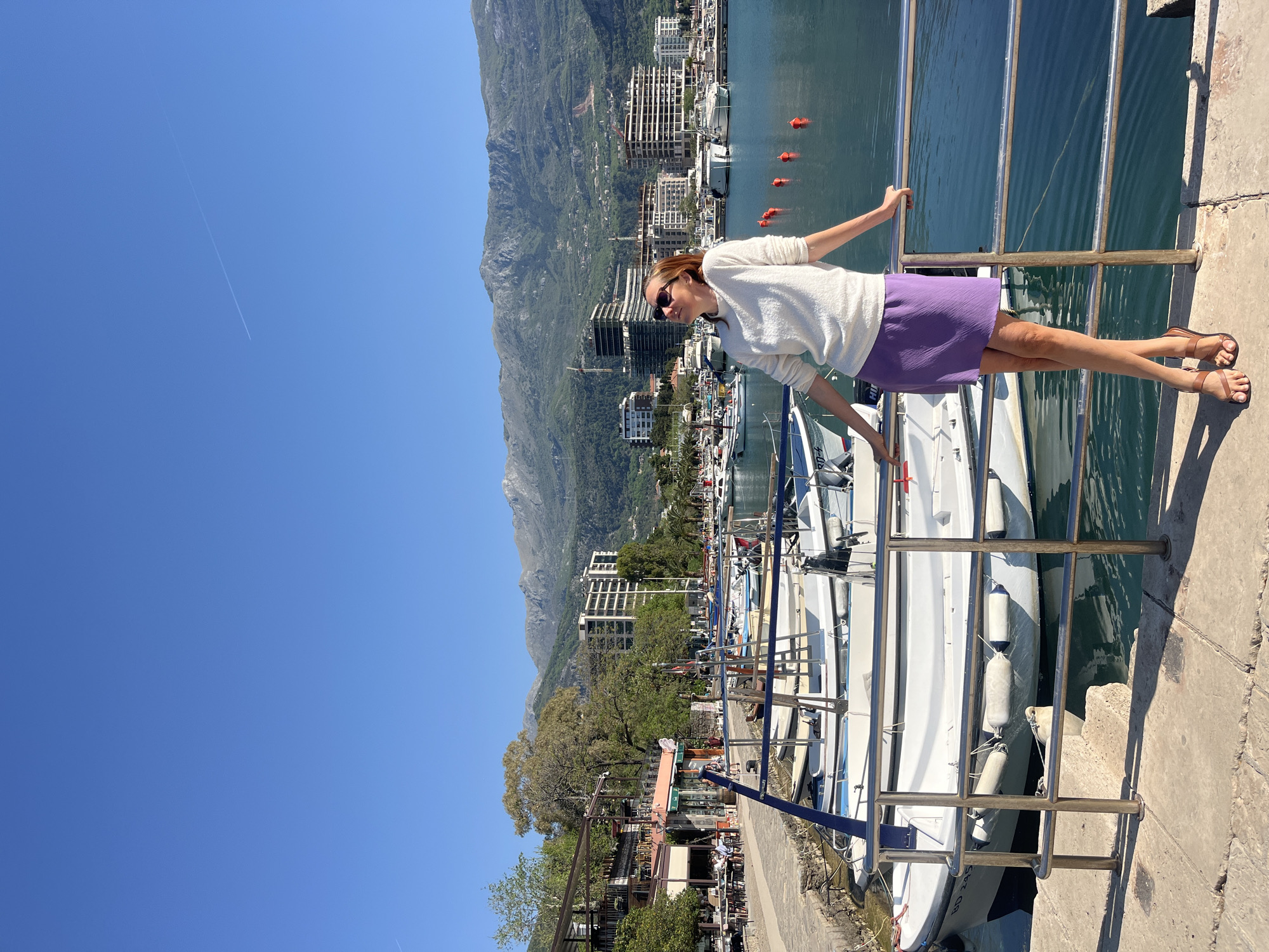 A woman stands adjacent to a railing separating a pedestrian walkway and a waterway. She wears sunglasses, a light-colored sweater, and a purple skirt. She leans against the railing, facing the background of the image.
The background includes a mountain range behind multiple buildings of varying sizes. In the waterway are several boats and floating orange buoys.
The scene takes place in Budva, Montenegro.