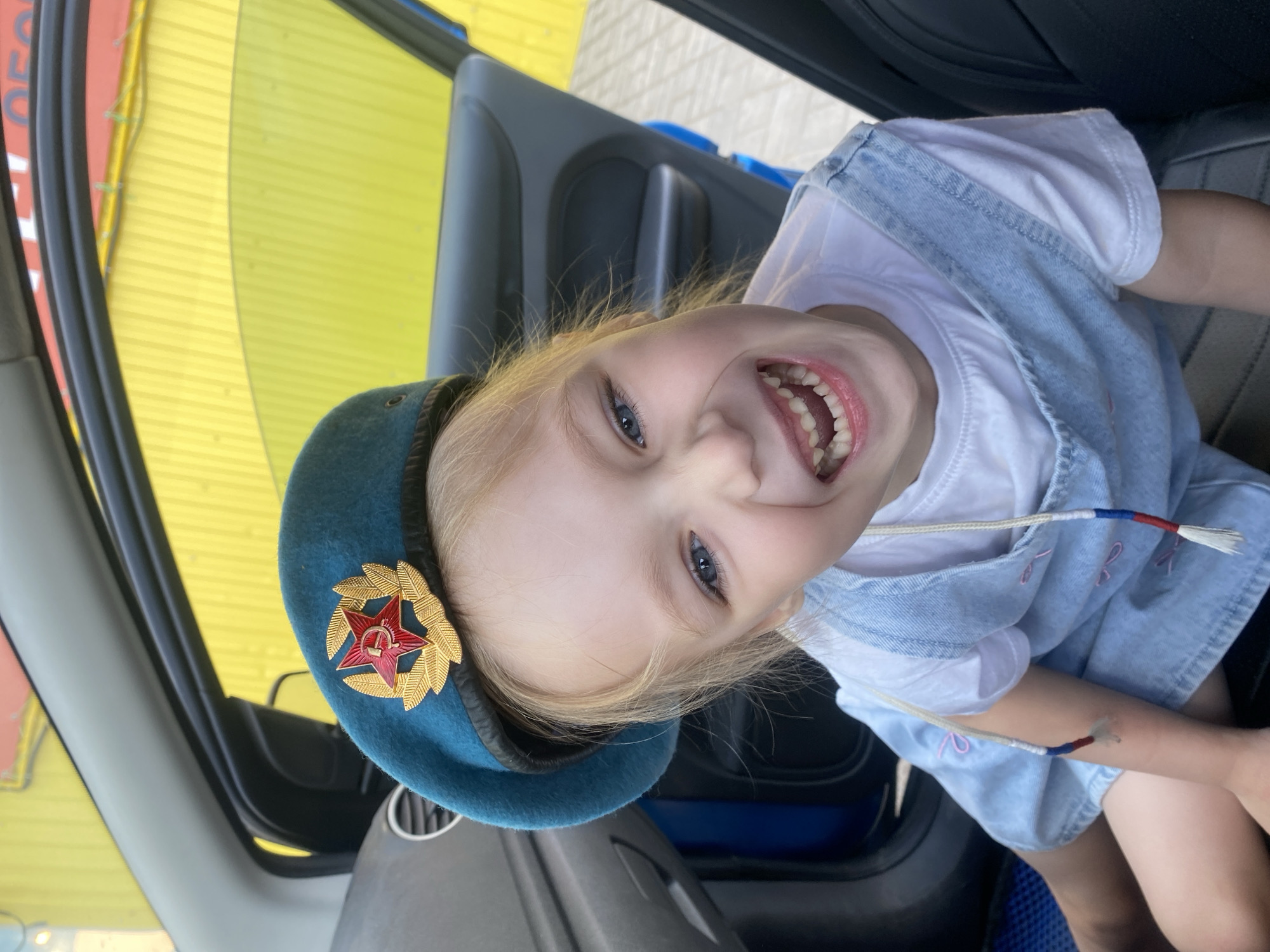 A young, fair-skinned, blonde-haired child with blue eyes is smiling broadly while sitting inside a vehicle. The child wears a white t-shirt and a light denim dress. On the child's head is a blue beret, adorned with a red star emblem surrounded by gold laurel leaves. A decorative white cord with red and blue accents hangs from the beret and drapes across the child's dress. The vehicle interior consists of dark grey elements and includes a section of the car door with a handle. Outside the vehicle, a bright yellow building exterior is visible. This image was captured in the city of Chaikovsky, Russia.