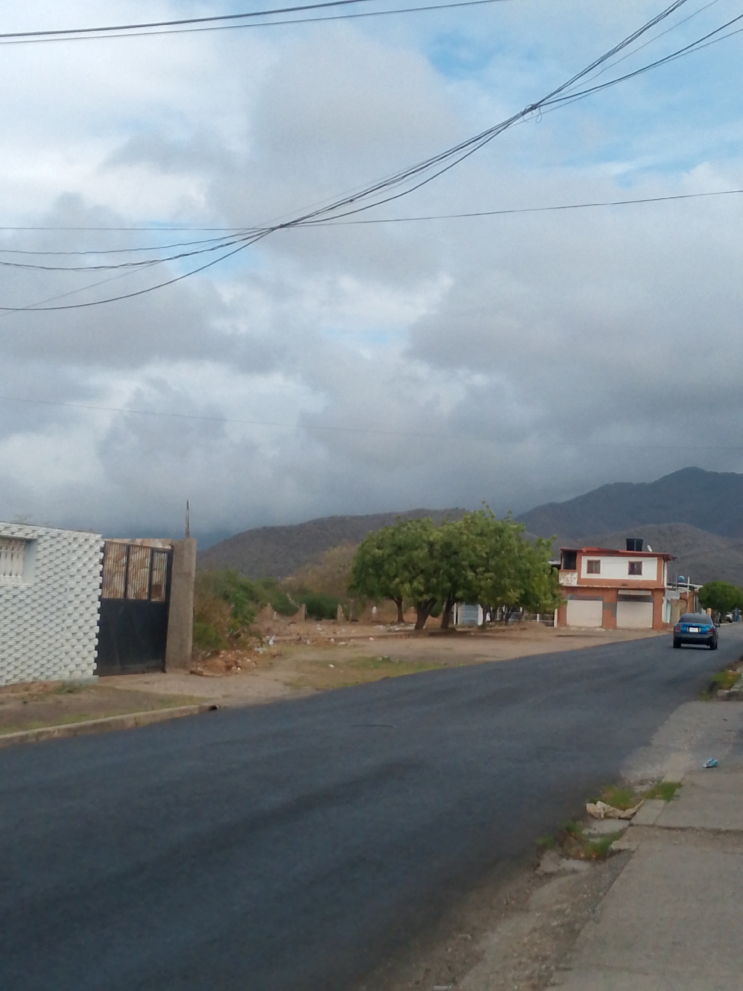The image presents a street view in Juan Griego, Venezuela, under an overcast sky. In the distance, rolling hills are visible. A two-story building with a white and orange facade stands on the right side of the street. A blue car is driving away from the camera. A cluster of trees is situated near the building. The road is asphalt, and a sidewalk runs along the right edge of the image. Power lines stretch across the top of the frame. On the left is a building with a white brick facade and a black gate. The area between the building and the road appears to be dirt and sparse vegetation. There is some visible trash scattered near the sidewalk.