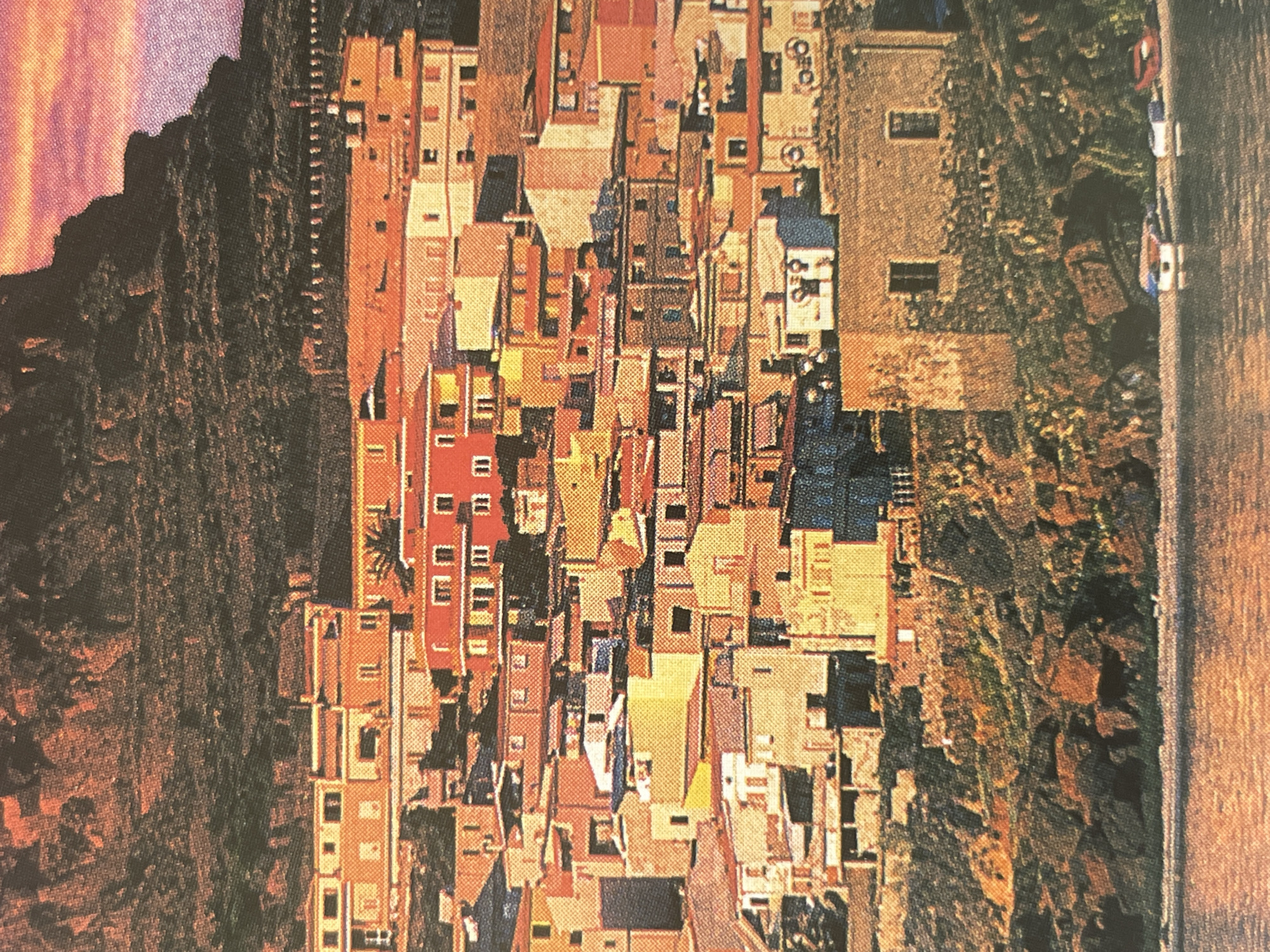 The image depicts a Mediterranean coastal town with densely packed, colorful buildings clinging to a hillside. The architecture is characterized by flat roofs, numerous windows, and a mix of pastel and vibrant hues, including orange, yellow, and red. The town appears to be situated on a rocky incline that slopes down towards a body of water, likely the sea or a large lake. The water in the foreground is calm and reflects the warm tones of the sky and buildings. Several small boats are visible pulled up on the shore. In the background, a dark, textured mountainside looms over the town, with a hint of pinkish-purple sky visible at the very top, suggesting either dawn or dusk. There are no people visible in the image, and the scene is devoid of any modern infrastructure beyond the boats. This scene is not located in the city of Sofia, Bulgaria.