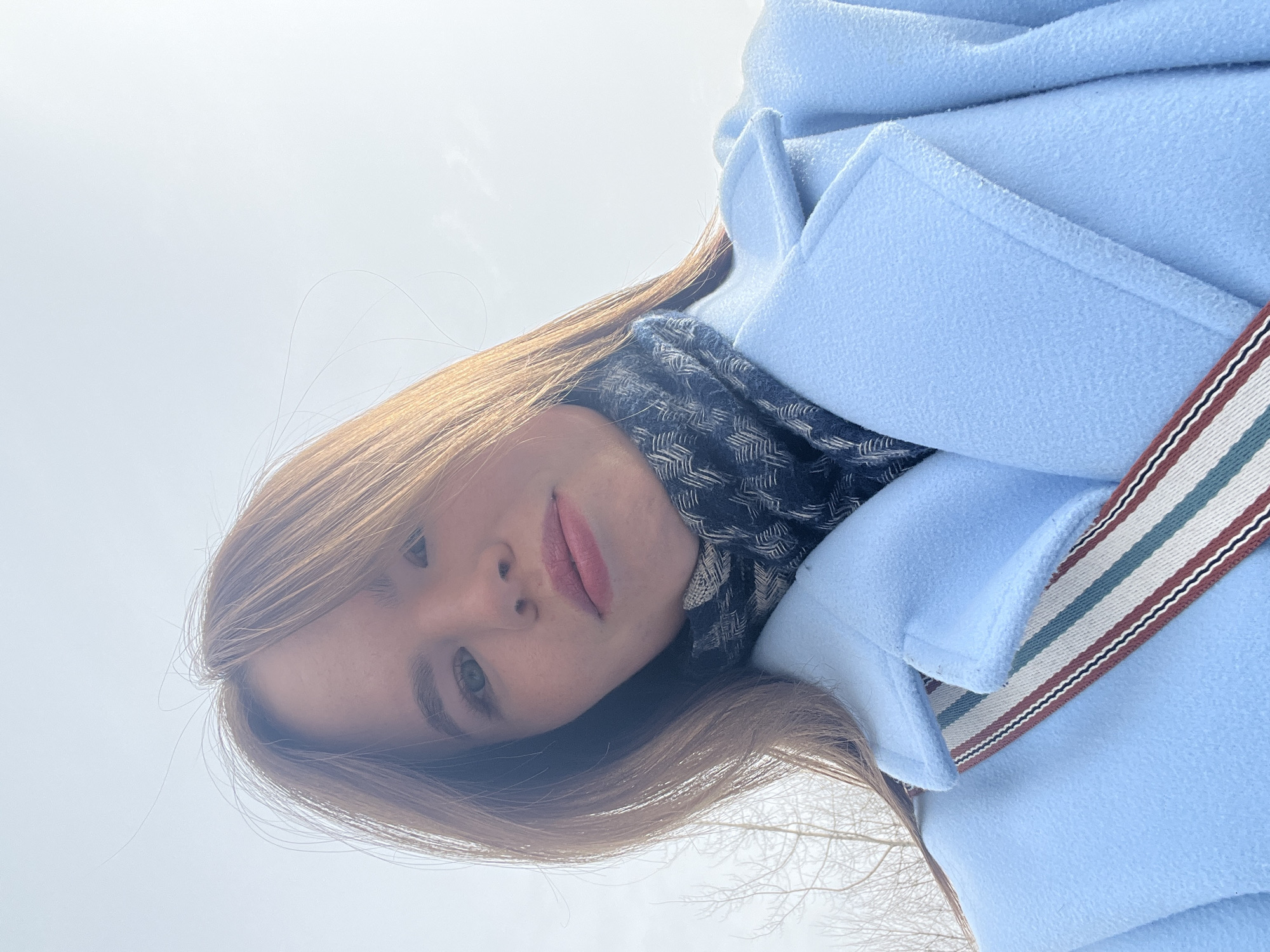 A young adult female is depicted in a low-angle selfie, looking directly at the camera. She possesses light brown hair, green eyes, and is wearing pink lipstick. Her attire includes a light blue coat with a wide collar and a dark scarf patterned with a light-colored geometric design (possibly herringbone or chevron) around her neck. A multi-colored striped strap, featuring white, red, green, and dark blue bands, crosses her chest diagonally. The background consists of a bright, overcast sky with the bare branches of deciduous trees visible on the left side, indicating an outdoor setting during a colder season. The precise location within the city of Chaikovsky, Russia, cannot be visually determined from the image.