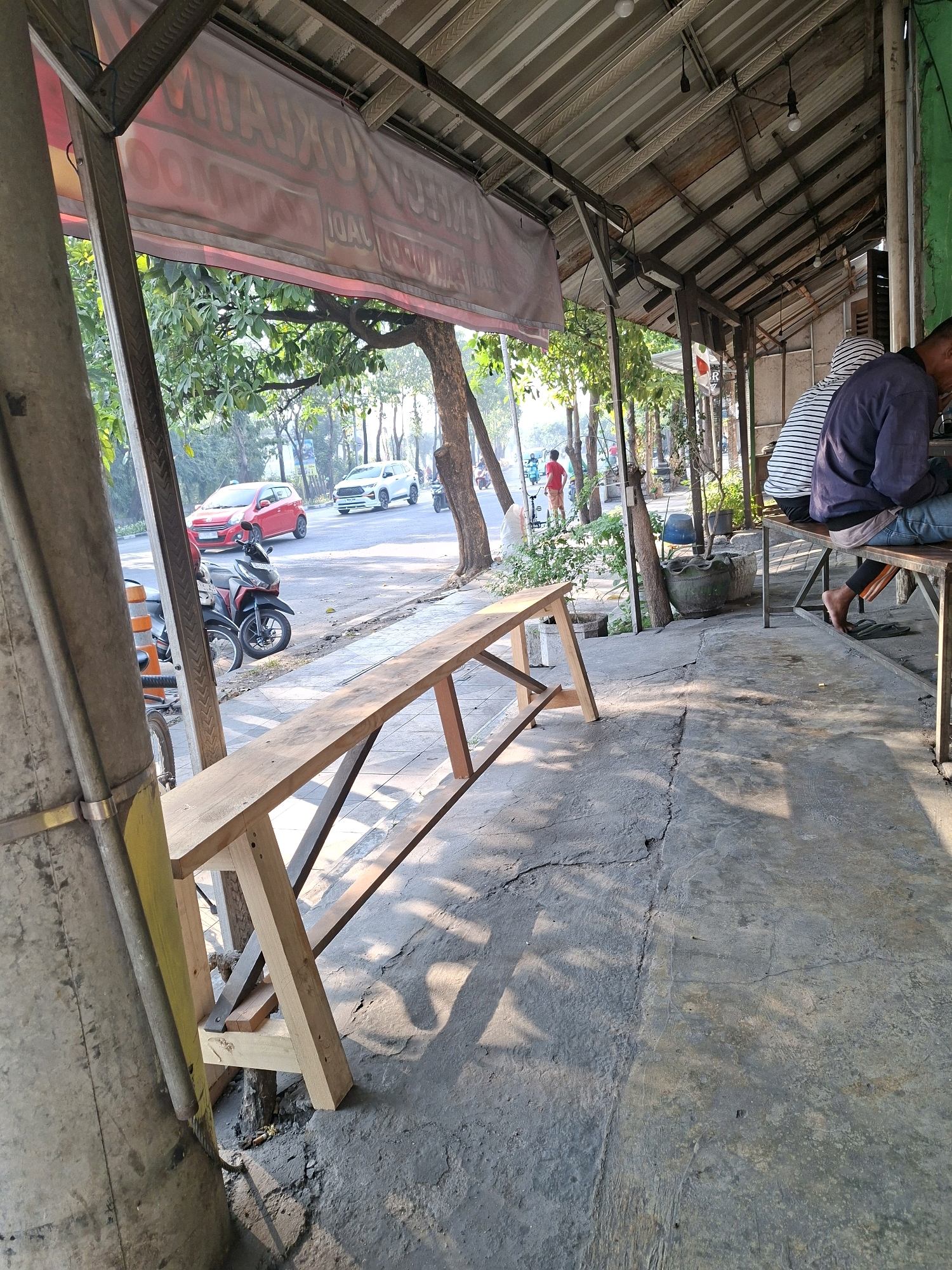 A roadside warung (small shop) in Surabaya, Indonesia, is captured in a wide shot. The foreground features a newly constructed wooden bench, its light-colored planks contrasting with the cracked concrete ground. Sunlight casts long shadows across the pavement, dappling the scene. A large concrete pillar stands to the left, partially obscuring the view of a street with moderate traffic. A red car and a white SUV are visible, along with several motorcycles parked on the side.
Behind the bench, the warung's structure is evident, with a corrugated metal roof supported by wooden beams. A translucent banner with red lettering, possibly advertising food or drinks, is hung across the front, partially visible and slightly distorted. Beyond the warung, lush green trees line the street, creating a verdant backdrop. Two individuals are seated on a longer, more weathered bench within the warung's shaded area. One person, wearing a dark long-sleeved shirt and jeans, is facing away from the camera, with their bare feet visible. The other, wearing a striped shirt, is also seated, their back mostly to the viewer. A few light bulbs hang from the ceiling, illuminating the interior. The overall atmosphere is that of a casual, everyday scene in an urban Indonesian setting.