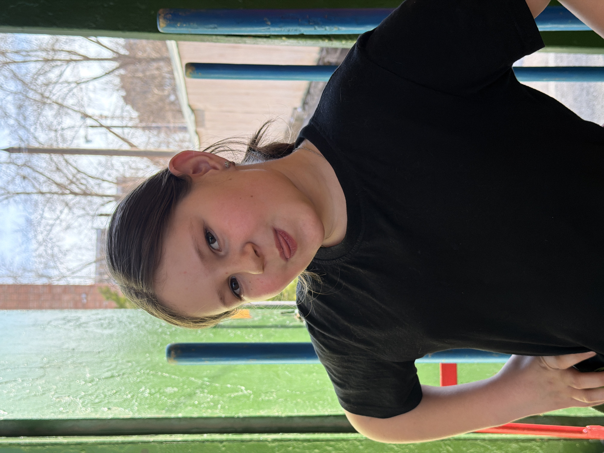 A girl wearing a black shirt is looking at the camera. She is outside on what appears to be a playground structure with green and blue bars. In the background are trees without leaves, and a brick building. It is not possible to determine the location is in Izhevsk, Russia from the image.