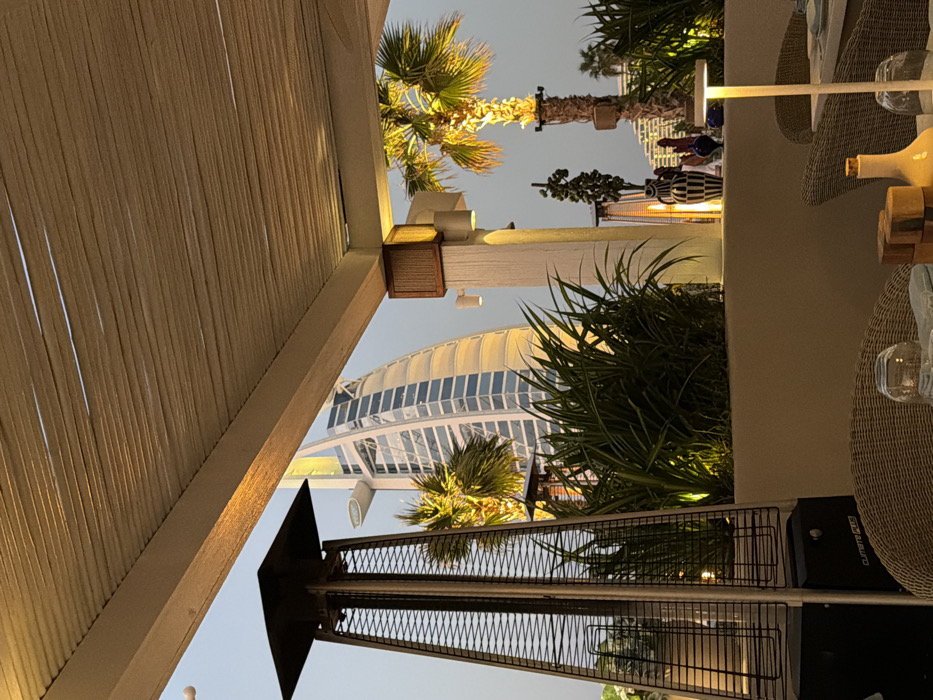 This image depicts an outdoor dining or lounge area in Dubai, United Arab Emirates, offering a view of the Burj Al Arab hotel. In the immediate foreground, a beige rattan chair is partially visible, alongside a tabletop with a clear glass, a light-colored bottle, and several stacked wooden blocks. To the left, a tall, dark-toned patio heater, featuring a pyramid-shaped mesh cage, is prominent. The midground contains a long, light-colored planter box filled with lush green foliage, illuminated from within. A stout white structural pillar with multiple integrated sconce lights stands behind the planter. To the right, a large palm tree with bright yellow-green fronds is illuminated from below, its trunk visible. Decorative objects, including a tall black and white striped vase, are placed on the planter's edge. In the background, the iconic sail-shaped structure of the Burj Al Arab, a luxury hotel, stands against a pale twilight sky, its reflective glass facade catching ambient light. No individuals are present in the scene. The area is illuminated by various artificial light sources, including the patio heater's glow, pillar lights, and a small, white portable table lamp on the rightmost table.