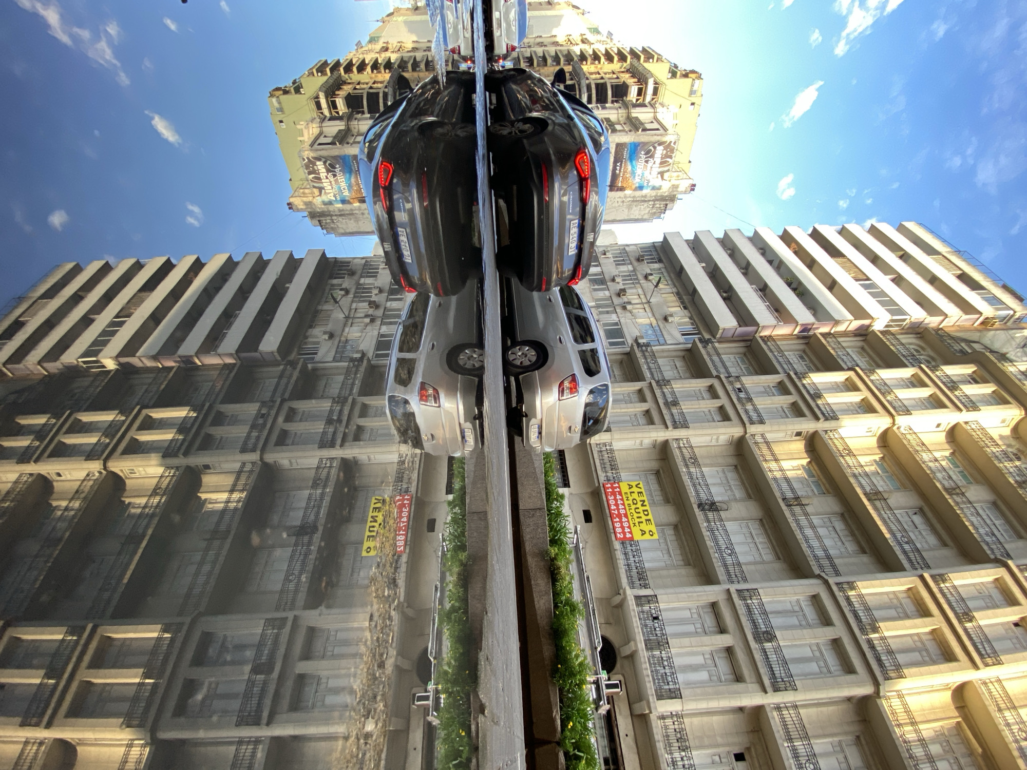 The image is a reflection of a street scene in Buenos Aires, Argentina, captured in a glass surface. A silver Chevrolet Corsa and a dark-colored Fiat Cronos are parked side-by-side. The reflection also shows adjacent buildings with fire escapes and advertising signage. A clear, blue sky with scattered clouds is visible. The signs are advertising properties for sale or rent.