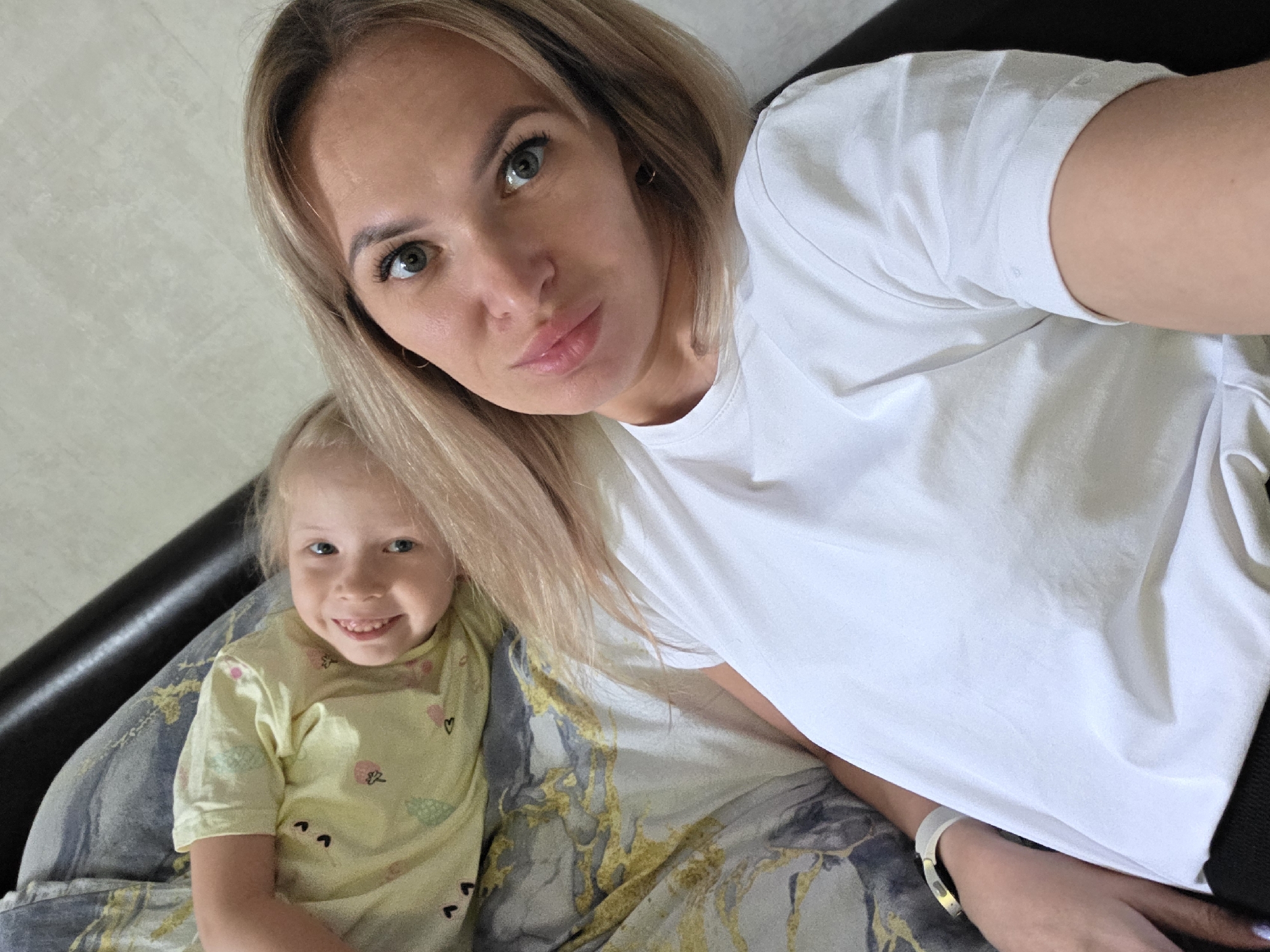 The image shows a selfie of two people, a woman and a young child, lying on a bed. The woman is in the foreground, with her face close to the camera. She has blonde hair and is wearing a white t-shirt. The child, who appears to be a little girl, is sitting next to her, slightly behind and to the left. She has blonde hair and is wearing a yellow t-shirt with a pattern of small shapes. Both subjects are looking at the camera, and the woman has a slight smile. The child is smiling widely, showing her teeth.
The background consists of a bed with patterned bedding, featuring abstract designs in shades of gray, blue, and gold. To the left, a dark object, likely the headboard of the bed or a pillow, is visible. The lighting suggests it could be daytime, possibly indoors in a bedroom. The overall mood appears relaxed and intimate, typical of a family moment. There is no visible text in the image. The location context provided is Chaikovsky, Russia.