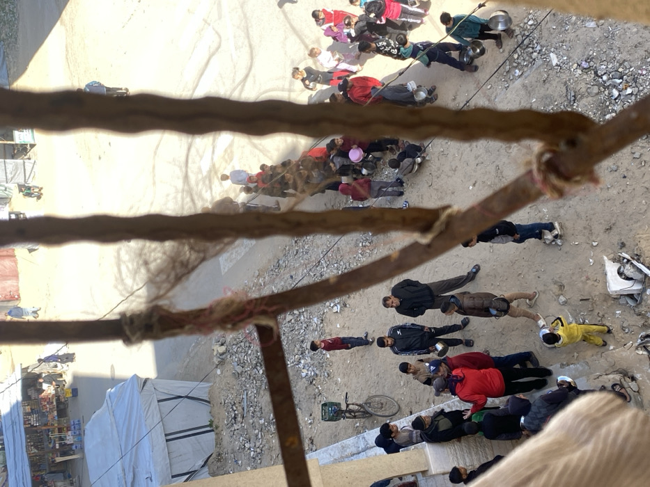 The image, captured from an elevated vantage point looking down, depicts an outdoor gathering in Khan Yunis, Palestinian Territory, partially obscured by three blurred, vertical wooden bars wrapped with twine in the foreground. The ground consists of uneven packed dirt, sand, and scattered rubble, stones, and debris.
To the immediate left, a light-colored building wall is visible, and further back, a large white tarp tent stands. In the background, a dirt road leads to what appears to be a market area with several temporary stalls constructed from corrugated metal sheets and other materials, displaying various goods. The scene is brightly lit by daylight, casting noticeable shadows.
Approximately 40-50 individuals are present, predominantly children, alongside several adults. On the left side, near the building, a group of adult males is engaged in conversation; one man in a red jacket is actively gesturing. A bicycle with a green crate on its rear rack leans against the building wall. Towards the center and right, a large group, primarily children, are gathered in an informal line or cluster. Many hold empty metal bowls, pots, or plastic containers, suggesting an organized distribution of items. A child in a bright yellow tracksuit in the lower center holds a blue container. The individuals wear various casual and winter clothing, including jackets and hoodies. Several adults are interspersed among the children. Individuals are also visible in the distant background near the market stalls.