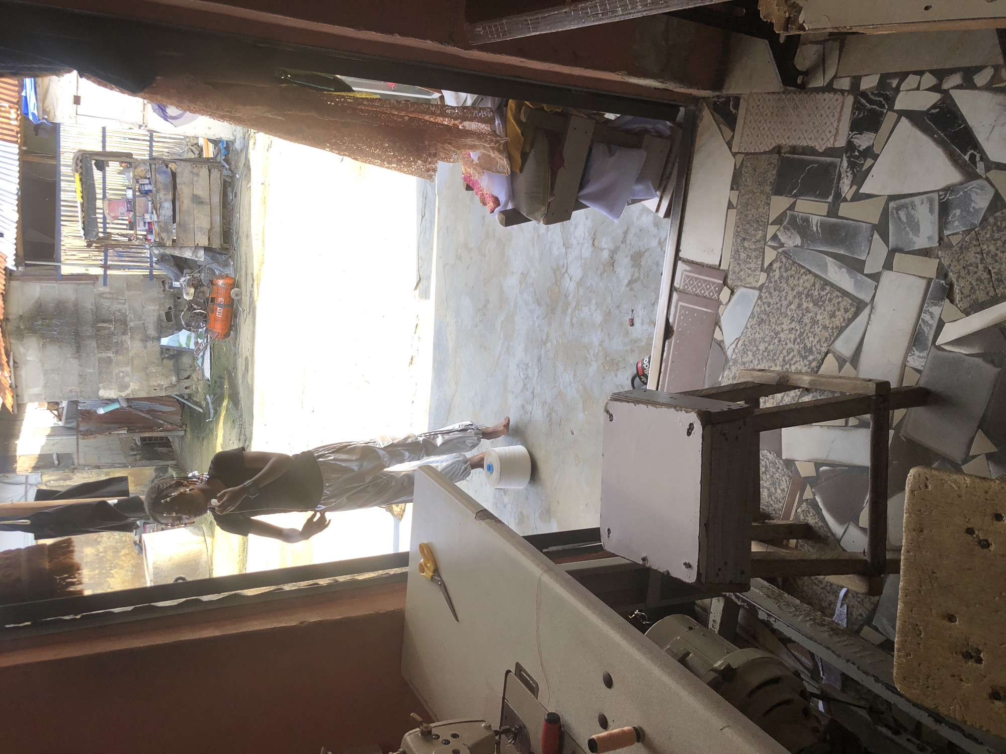 The image depicts the interior of a tailor's shop in Ado, Nigeria, looking outwards towards an open doorway. The shop is cluttered with sewing equipment, including a sewing machine on the left and a stool in the foreground. A large spool of white thread is on the floor behind the stool. In the doorway, a young person wearing a dark t-shirt and shiny pants stands with their hand to their mouth, appearing to be eating or chewing something. The background outside the shop shows a dusty, unpaved area with corrugated metal structures, a cement well, and an orange air compressor. Sunlight illuminates the scene. The flooring inside the shop is a mosaic of broken tiles.