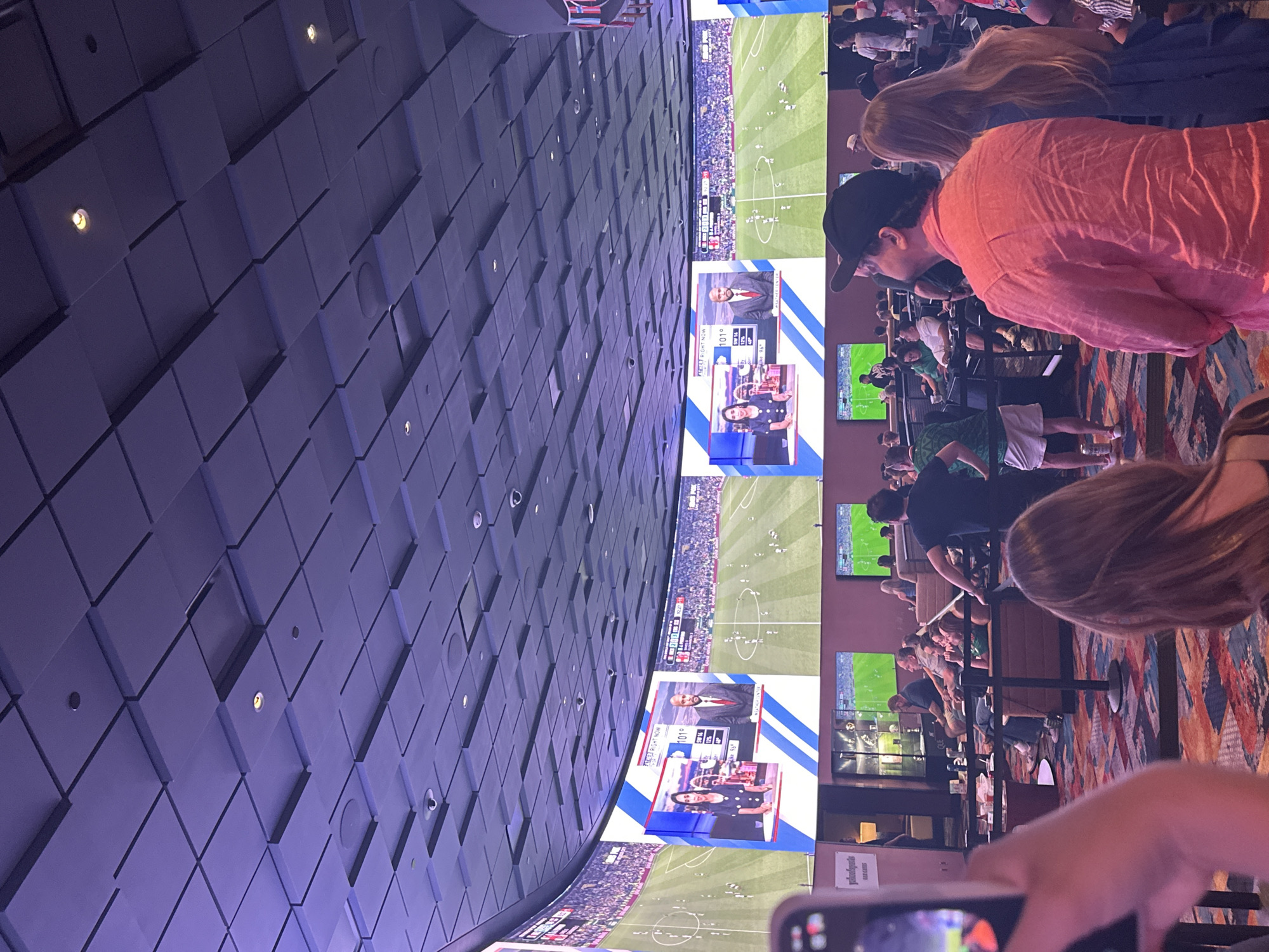 The image depicts a scene inside a sports betting venue in Las Vegas, Nevada, USA. The venue features a large, curved LED screen displaying a soccer match. In the foreground, several people are visible, some standing and others seated, watching the screen. One person in the foreground is holding a smartphone, presumably to take a photo or video. The ceiling of the venue is also visible, with a distinctive patterned design. The overall atmosphere appears to be one of casual entertainment and sports viewing.
