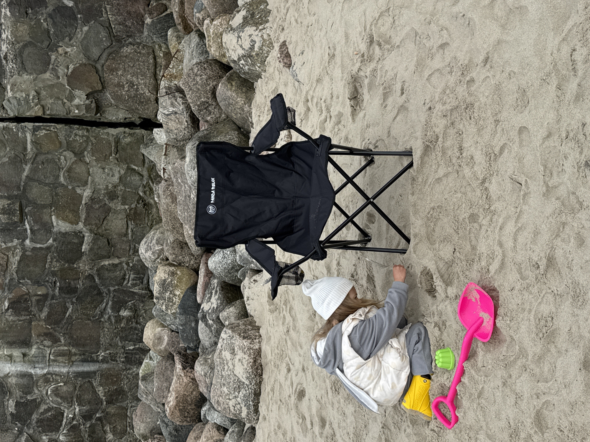A young child is sitting in the sand next to a black folding chair on a beach in Zelenogradsk, Russia. The child, wearing a white beanie, grey sweater, white vest, grey pants, and yellow Crocs, is playing in the sand. A pink shovel and a green toy are nearby. To the left of the chair are large grey rocks, possibly part of a breakwater or coastal defense structure. The sand is light-colored and textured with footprints and other markings.