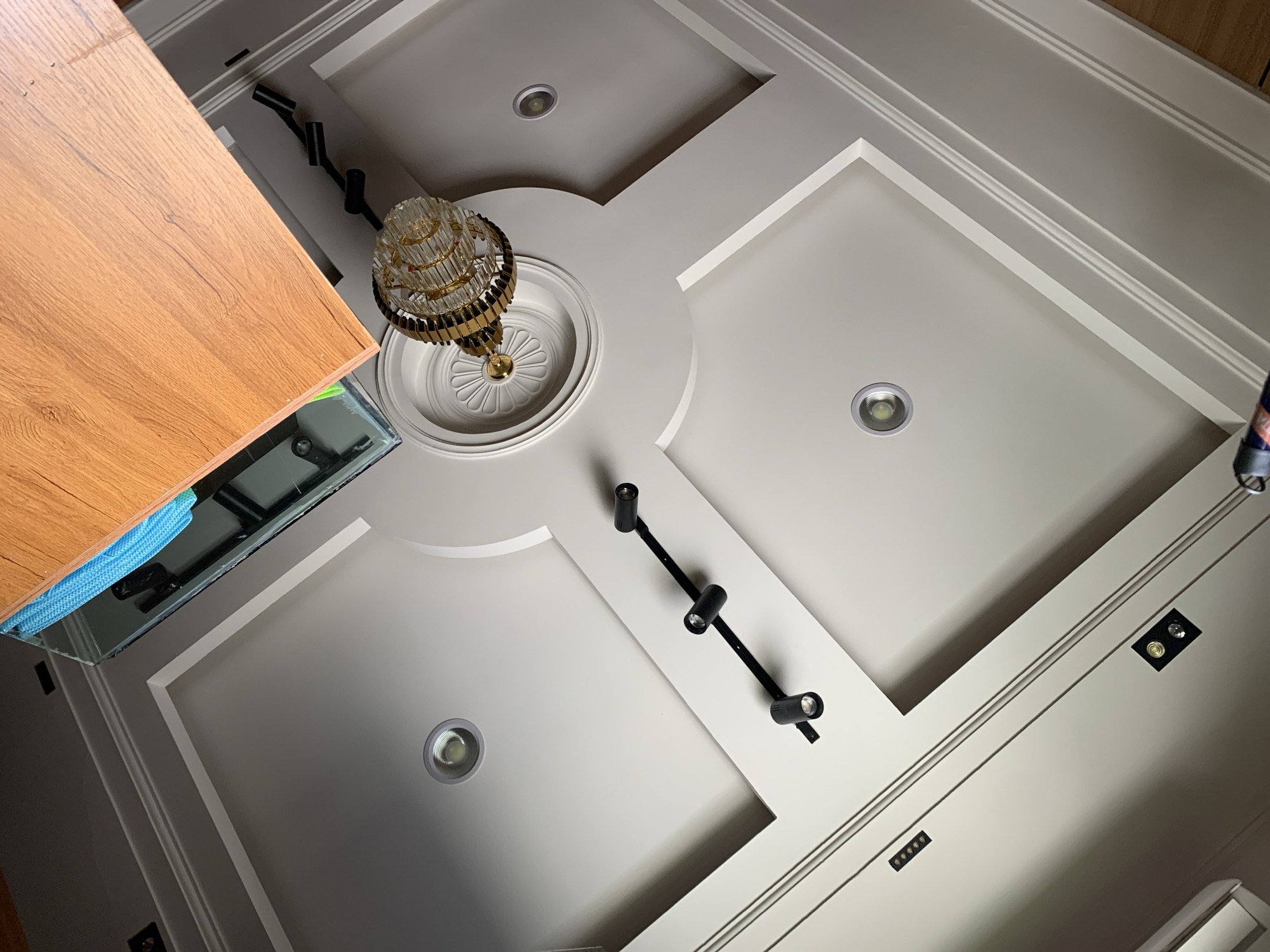 The image shows a ceiling with a wooden shelf and a chandelier. The ceiling is decorated with recessed lighting and molding. The shelf appears to be made of light-colored wood and has a glass edge. The chandelier is made of crystal and brass. The image was taken indoors, likely in a residential or commercial space. There is no indication of the specific location in Gura Top, Nigeria.