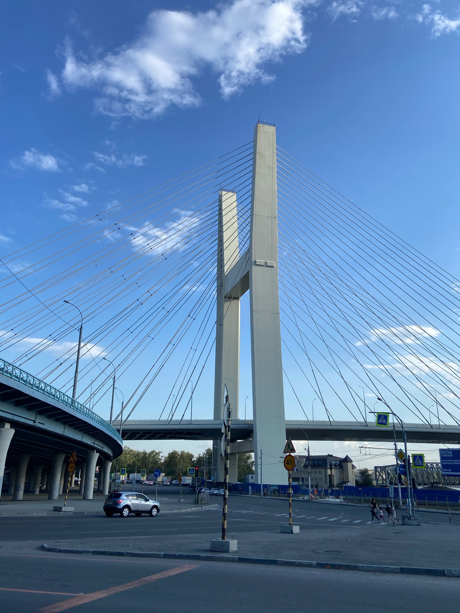 In Novosibirsk, Russia, a wide view captures the imposing presence of the Bugrinsky Bridge, a cable-stayed bridge, under a bright blue sky dotted with scattered white clouds. The two massive, white pylons of the bridge dominate the center of the frame, with a dense network of supporting cables fanning out from them. To the left, a curved elevated highway, supported by numerous concrete pillars, angles into the scene. The foreground shows a paved road with painted yellow lines, suggesting an intersection. A silver hatchback car is stopped in the middle of the road. Traffic signals, road signs, and streetlights are visible. In the background, beyond the elevated highway and the main bridge structure, a few buildings and trees are discernable. A pedestrian crossing is marked on the road in the mid-ground, with a few people walking across it. The overall scene depicts a dynamic urban landscape dominated by impressive infrastructure.