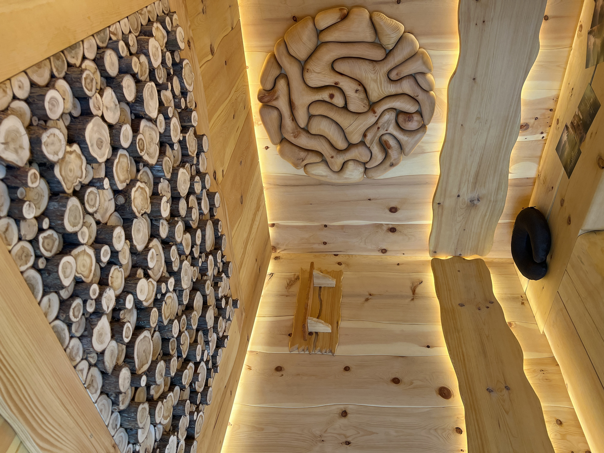 The image depicts the interior of a room constructed entirely of wood, likely a sauna or spa facility in Yakunino, Russia. The ceiling is adorned with a mosaic of circular wood slices of varying sizes, creating a natural, organic pattern. The walls are paneled with vertical wooden planks, and accent lighting runs along the top edges of the walls, casting a warm, yellow glow.
Dominating the right wall is a large, circular wooden sculpture with an intricate, maze-like design. On the left wall, a small, rustic wooden shelf is mounted. Built-in wooden benches line the lower portion of the room. On one of the benches, a black neck pillow rests, and several pieces of paper, possibly brochures or menus, are laid out. The overall aesthetic is rustic and natural, emphasizing the use of wood in both construction and decoration. There are no people visible in the image.