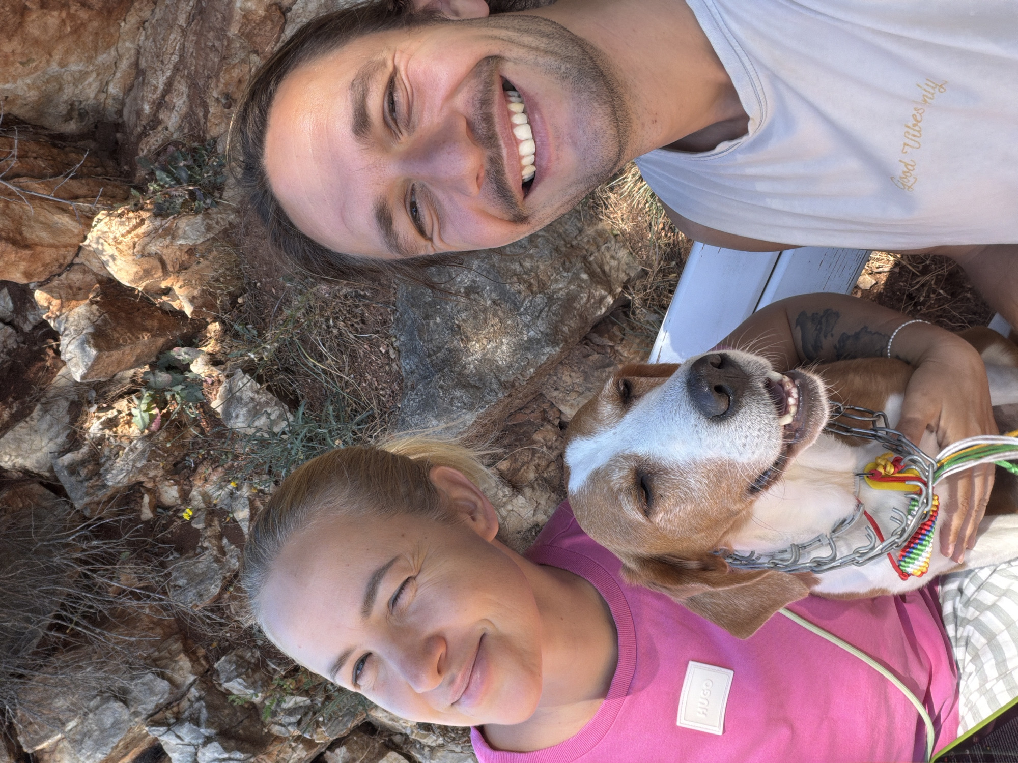 The image shows two adults and a dog lying on what appears to be a rocky surface, possibly in an outdoor setting. The man, on the upper right, is smiling with his teeth visible, wearing a white tank top with the inscription "Good Vibes Only", laying on the edge of a white bench. The woman, on the bottom left, has her eyes partially closed, as though she is smiling and squinting in bright light. She wears a pink t-shirt with a HUGO brand label on it. The dog, positioned between them, has brown and white fur and a distinctive metal prong collar adorned with colorful woven cords. The rocky surface is predominantly brown with some patches of green vegetation. The location is Mirište, Montenegro.