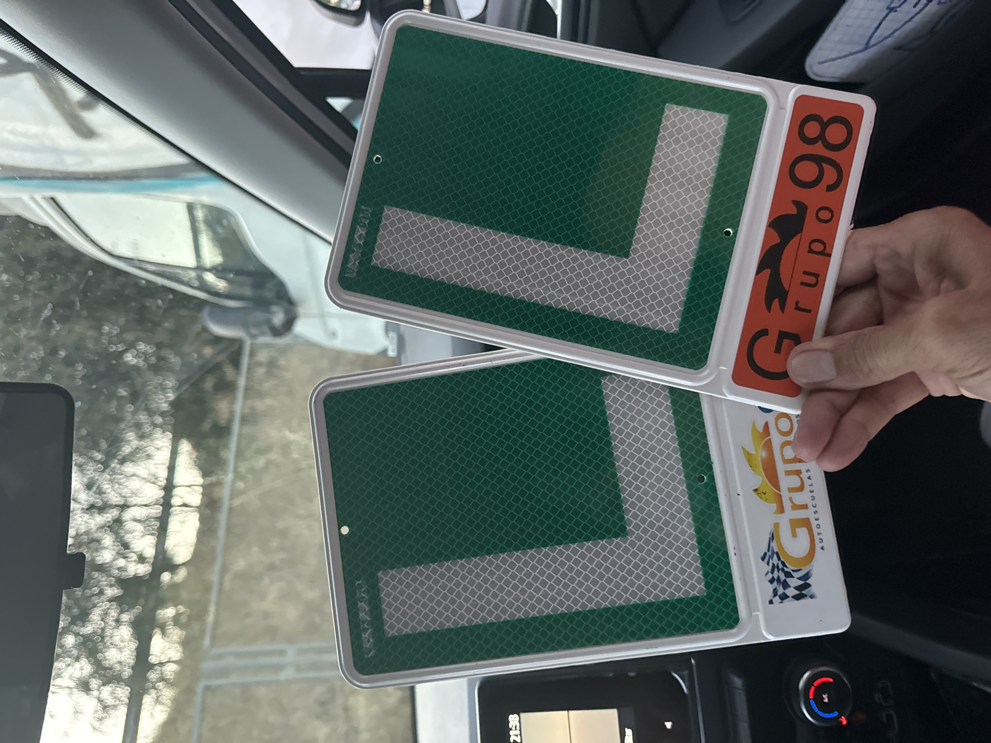 The image shows two green learner driver signs with reflective white 'L's inside, held by a person's hands. The signs are in the interior of a car, with a dashboard visible on the left. The background outside the car is blurred, showing trees and a white vehicle, suggesting an outdoor setting. The learner signs are branded with "Grupo 98 AUTOESCUELAS" and a checkered flag logo, indicating they are from a driving school. The person is likely in Murcia, Spain, as "AUTOESCUELAS" is a Spanish term for driving schools. The overall scene is that of a learner driver preparing to practice, with the car's interior and the learner signs being the focal points.