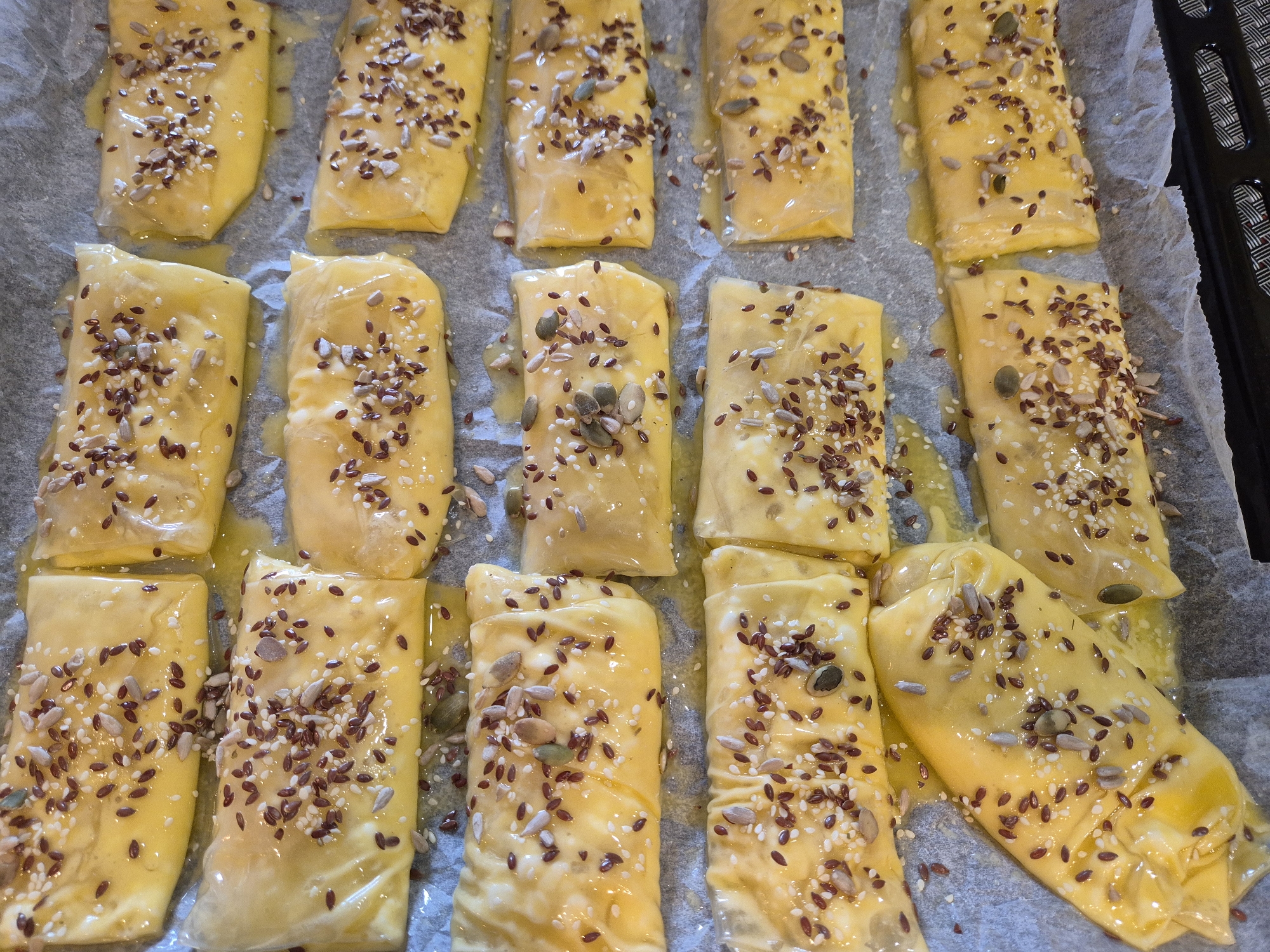 Captured from an overhead perspective, the image presents a close-up shot of a batch of pastries arranged on a baking sheet covered in parchment paper. The pastries, rectangular in shape, have a glistening yellow hue, likely due to a glaze or egg wash applied to their surface. Each pastry is sprinkled with a mix of seeds, including sunflower, sesame, and flax seeds, adding visual texture and presumably enhancing the flavor and nutritional content. The parchment paper exhibits creases and folds, indicating it has been used as a non-stick surface during the baking process. The background features a portion of a baking rack, suggesting the setting is a kitchen. The location (city and state) is not ascertainable from the provided image.