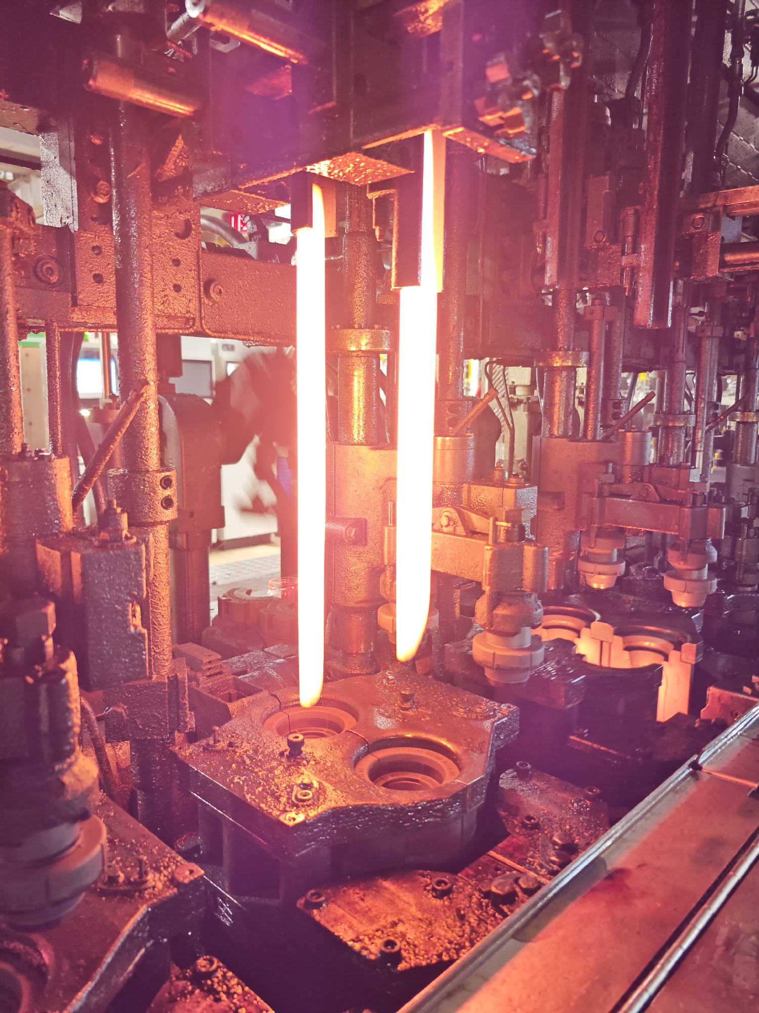 The image depicts the interior of a glass manufacturing facility, possibly located in Pleven, Bulgaria, given the prompt's request. The scene focuses on a complex piece of industrial machinery, heavily lubricated with oil or grease. Two intensely bright, elongated flames or streams of molten glass are centrally positioned, emerging from above and descending towards a dark, multi-chambered mold below. Various mechanical components – cylinders, rods, and joints – are visible, suggesting automated processes. There is a suggestion of a person in the blurred background in the scene. The environment is industrial and functional.