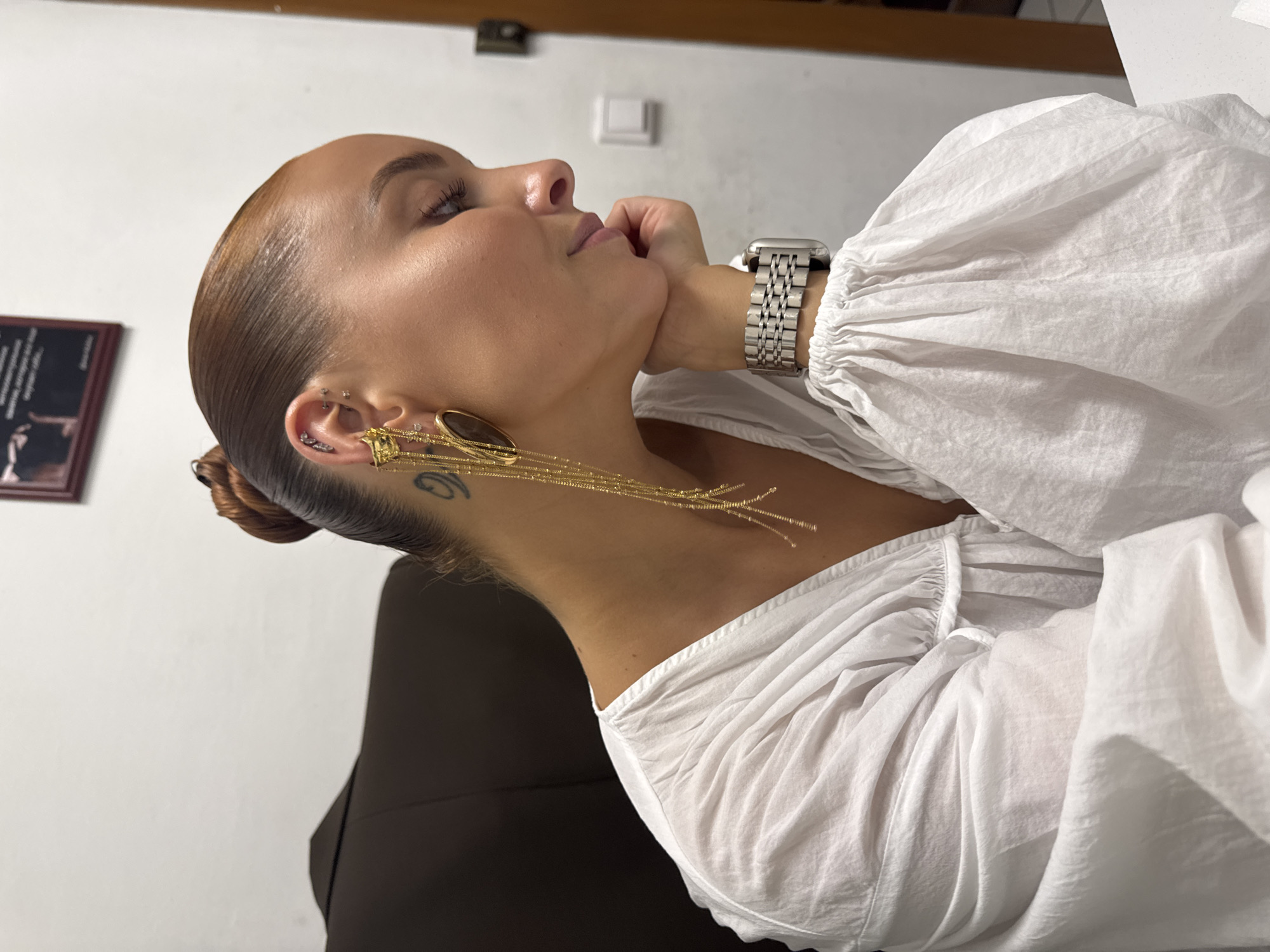 In a dimly lit room, a woman with her hair pulled back in a bun is captured in a side profile, resting her chin on her hand. She is wearing a white blouse with gathered sleeves and a silver watch. A long, golden earring dangles from her left ear, adorned with small beads and an oval-shaped pendant. A tattoo is visible on the back of her neck. The background consists of a white wall with a framed picture and a dark-colored chair. There is no specific indication of the location being in the city of Chaikovsky, Russia.