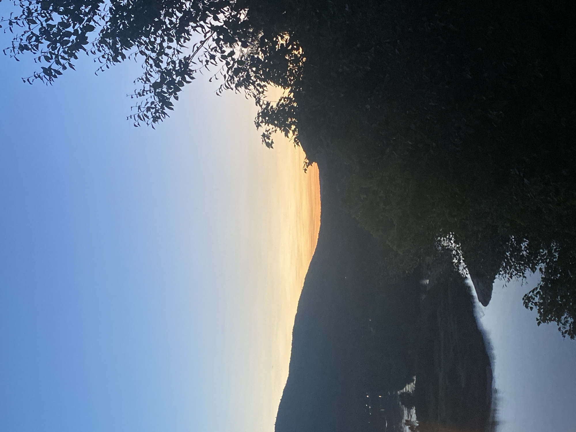 An outdoor photograph captures a serene twilight scene overlooking a body of water and rolling hills, likely in Saint-Gabriel-de-Valcartier, Canada. The sky transitions from a deep, clear blue at the top to a soft gradient of pale yellow and orange near the horizon, indicating the setting or rising sun. Silhouetted against the illuminated sky are the dark shapes of trees and a mountain range. In the foreground, the dark foliage of trees frames the right side of the image. Below the tree line, a wide river or lake curves through the landscape, its surface reflecting the dim light of the sky. On the far side of the water, the dark outline of a low-lying area with scattered lights suggests a settlement or distant buildings. The overall impression is one of tranquility and natural beauty as day transitions into night. There are no people or animals visible in the frame.